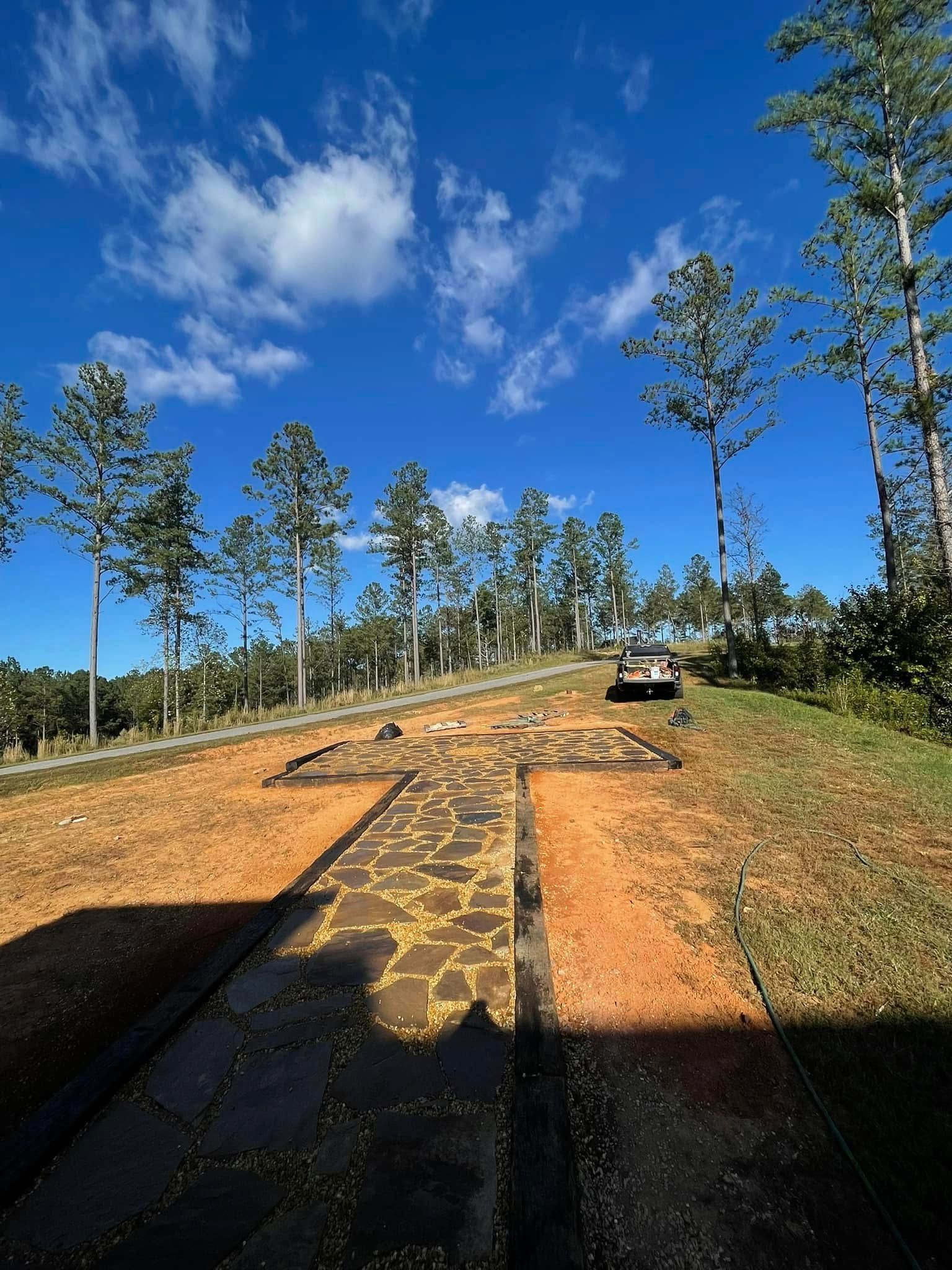 A stone walkway shaped like a cross leads toward a parked car on a grassy hill under a bright, sunny blue sky.