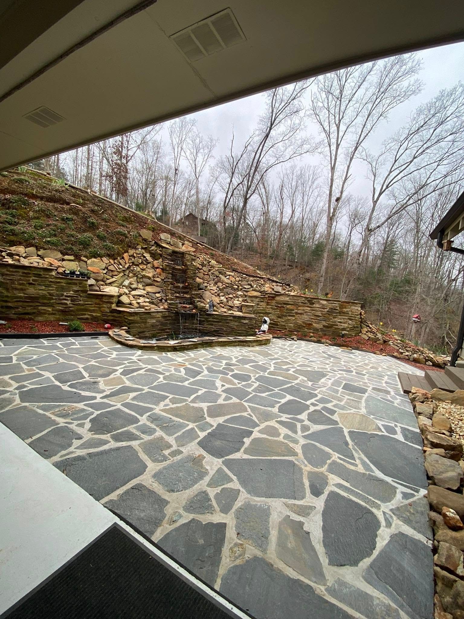 An outdoor patio featuring gray flagstone pavers, set against a stone retaining wall and a wooded hillside.