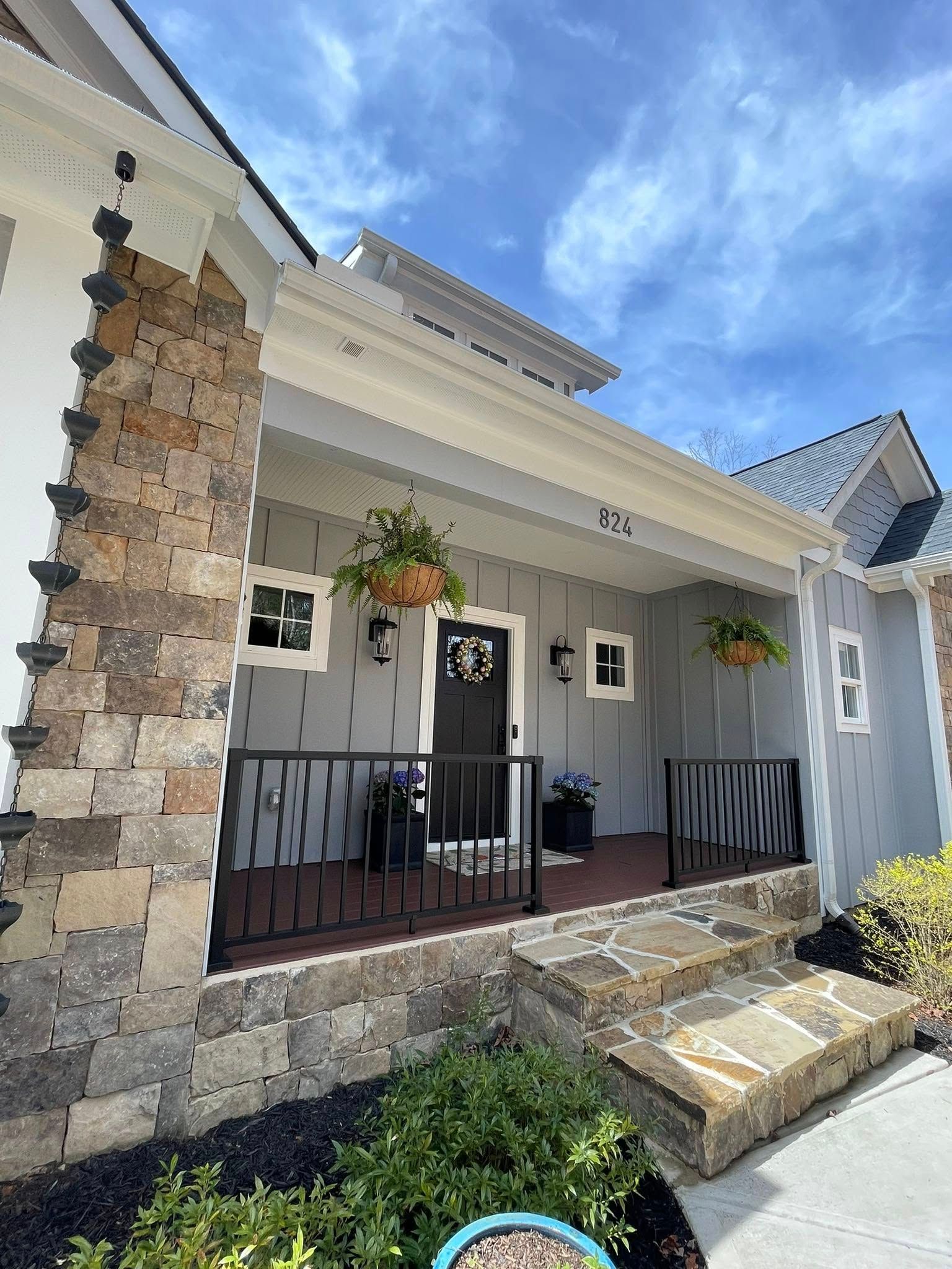 A house porch with gray siding, stone pillars, black railings, and a front door with a wreath under a blue sky.