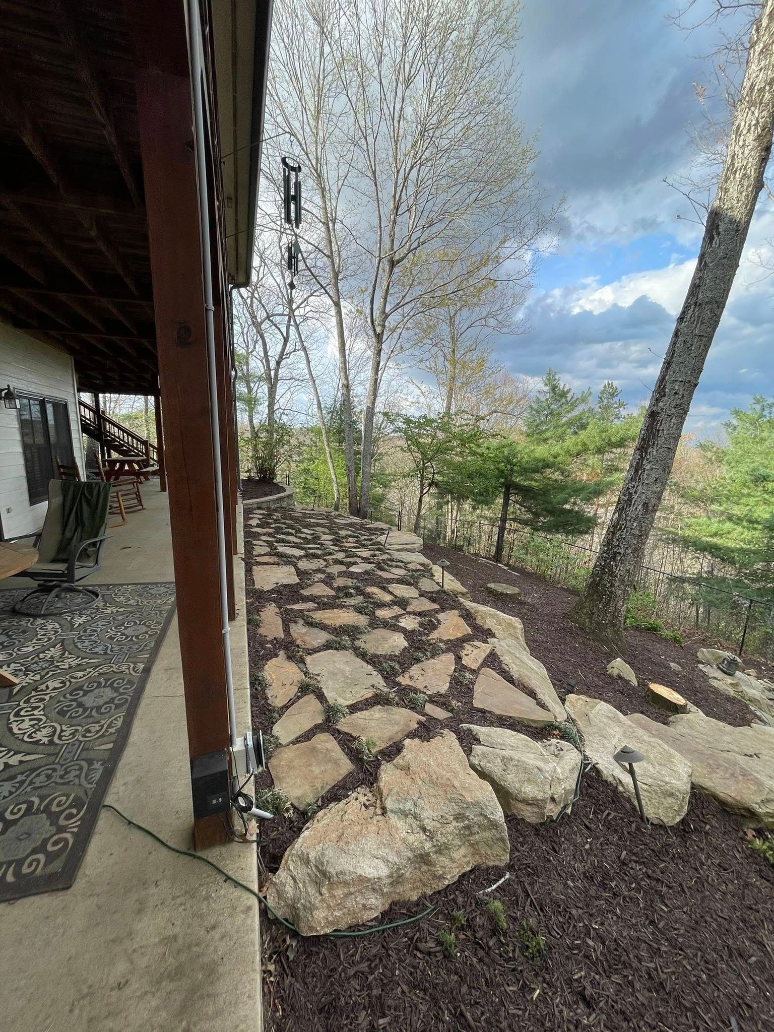 A stone walkway path runs along the edge of a house porch next to mulched garden beds and trees under a cloudy sky.