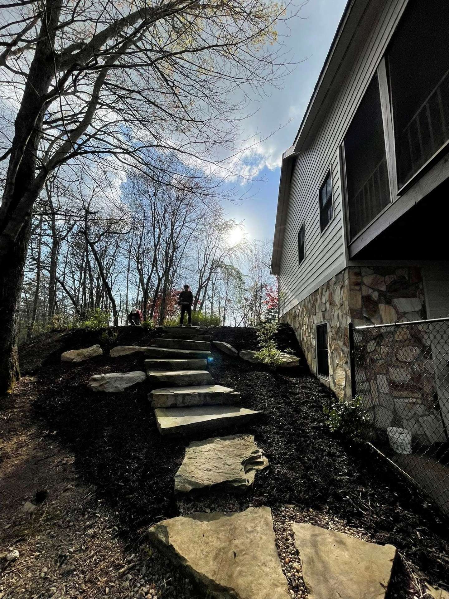 Stone steps lead up a mulched path toward a silhouetted figure under a bright sun, next to a multi-story house.