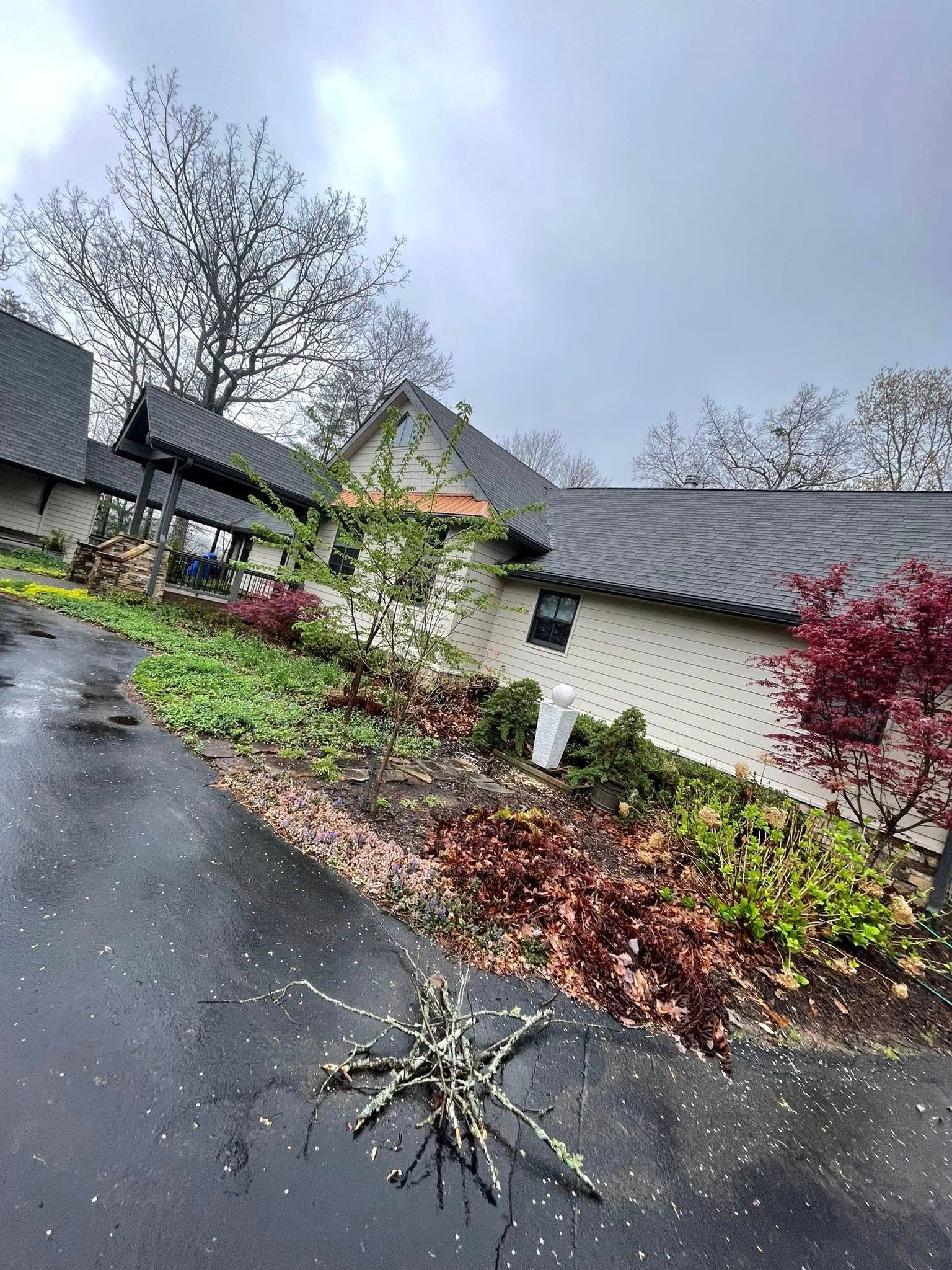 An asphalt driveway leads to a light-colored building with a dark shingled roof, surrounded by trees and landscaping.