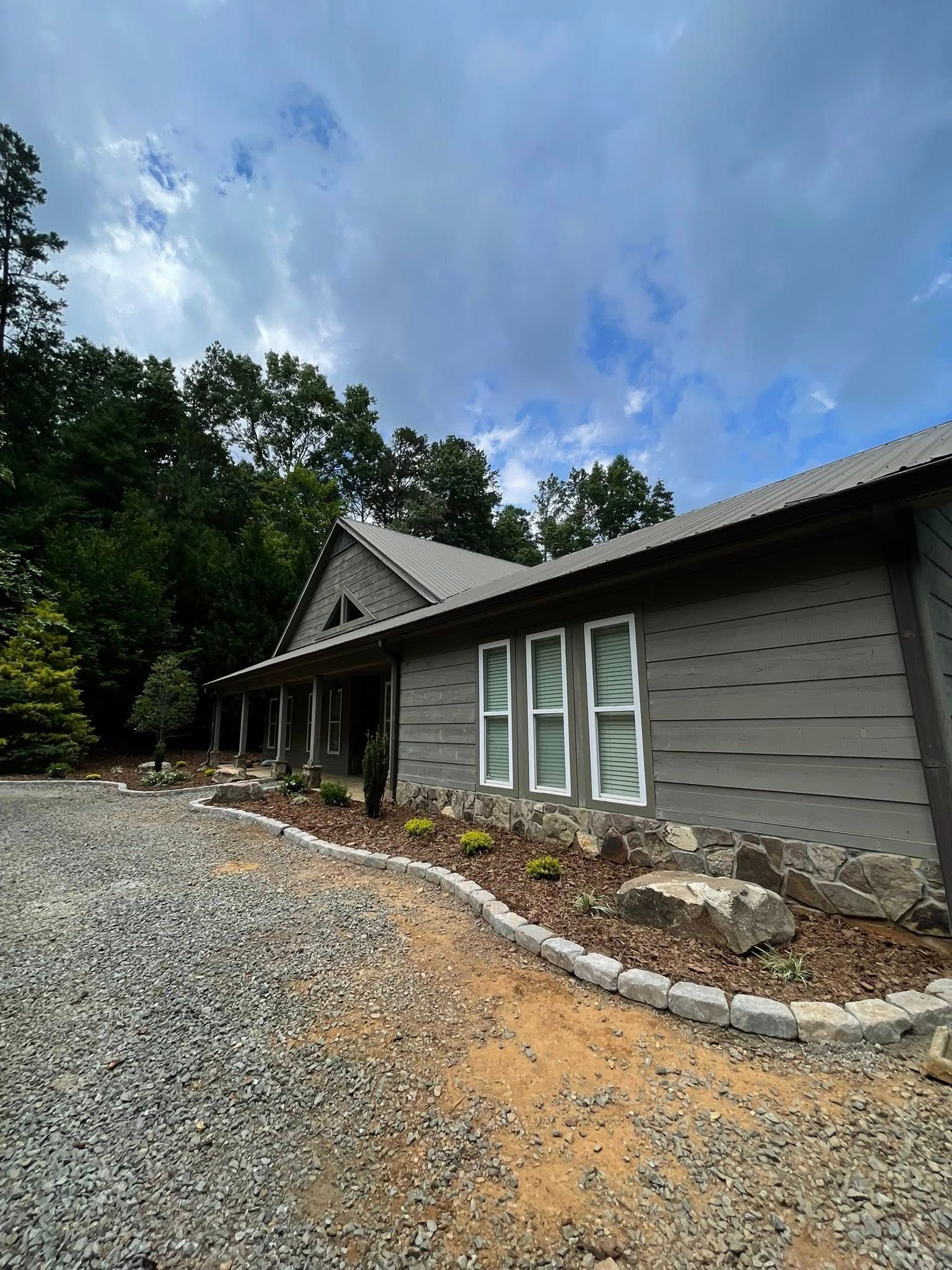 A gray, one-story cabin with a stone foundation, a metal roof, and a gravel driveway set against a backdrop of trees.