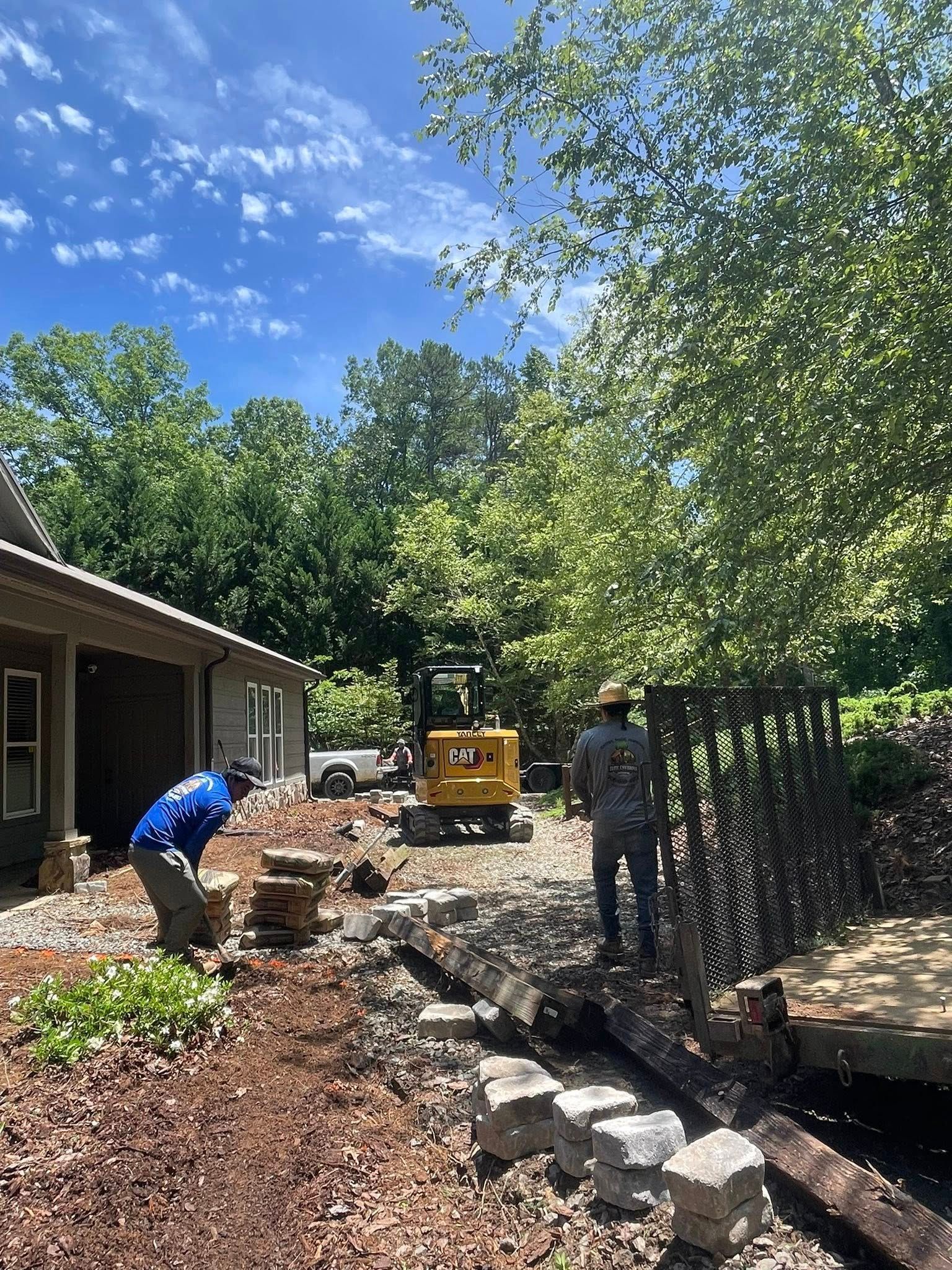 Two workers in a yard operate a small yellow excavator near a house with landscaping stones and trees on a sunny day.