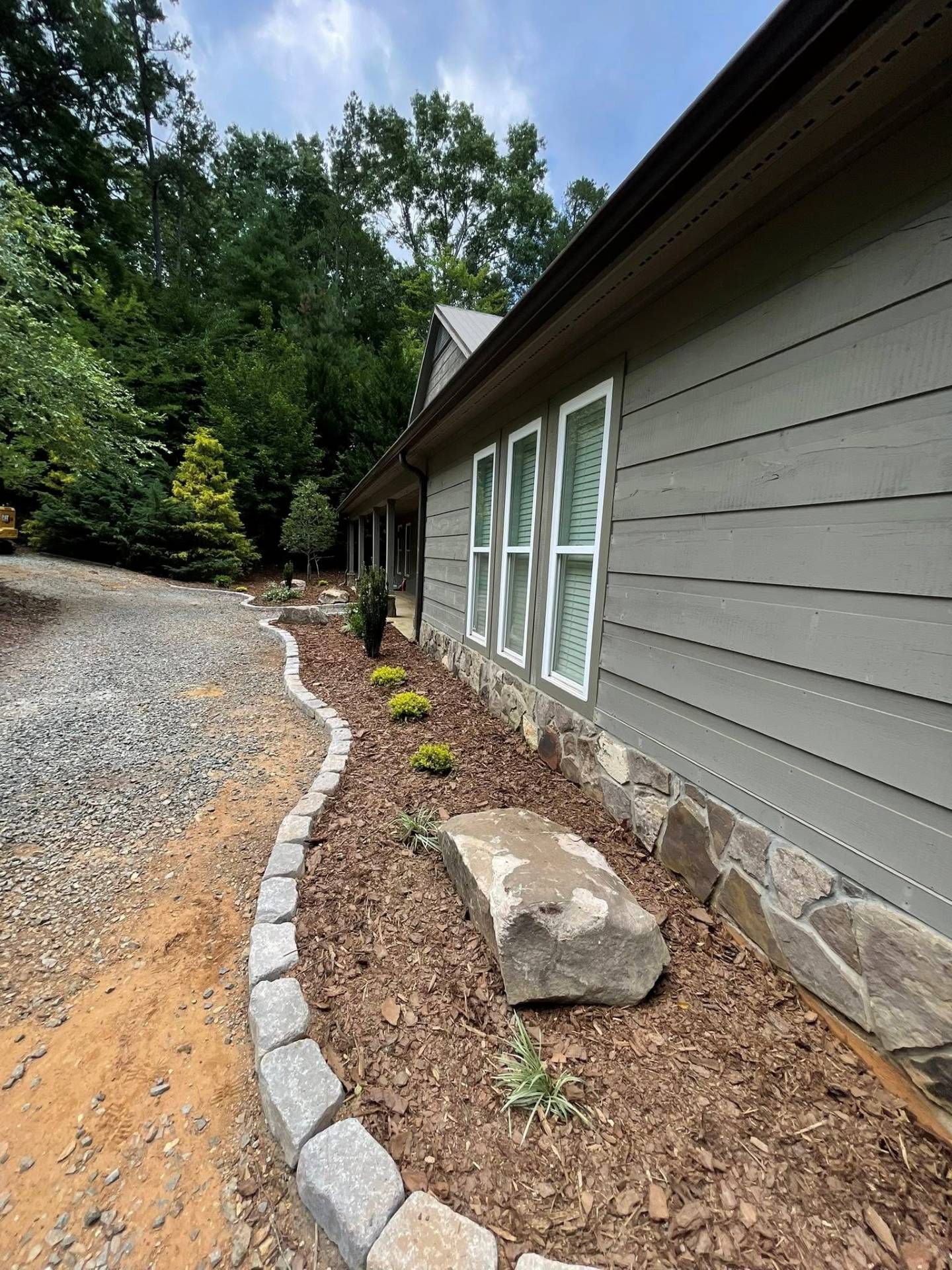 A gravel path borders a mulched garden bed with small plants and a large rock, alongside a house with gray siding.