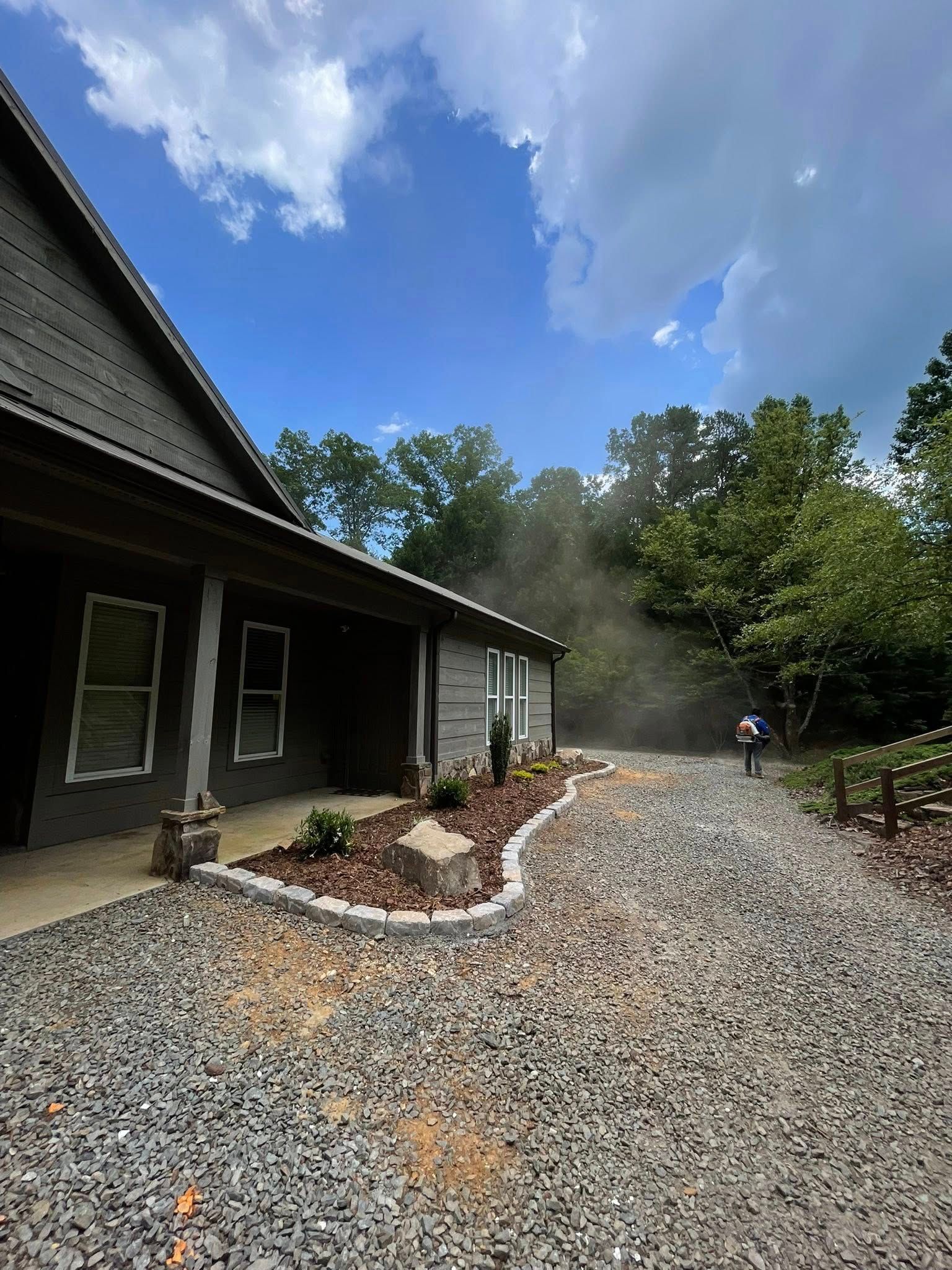 A gravel driveway leads to the side of a grey house with a porch and a small, stone-bordered garden bed under a sunny sky.