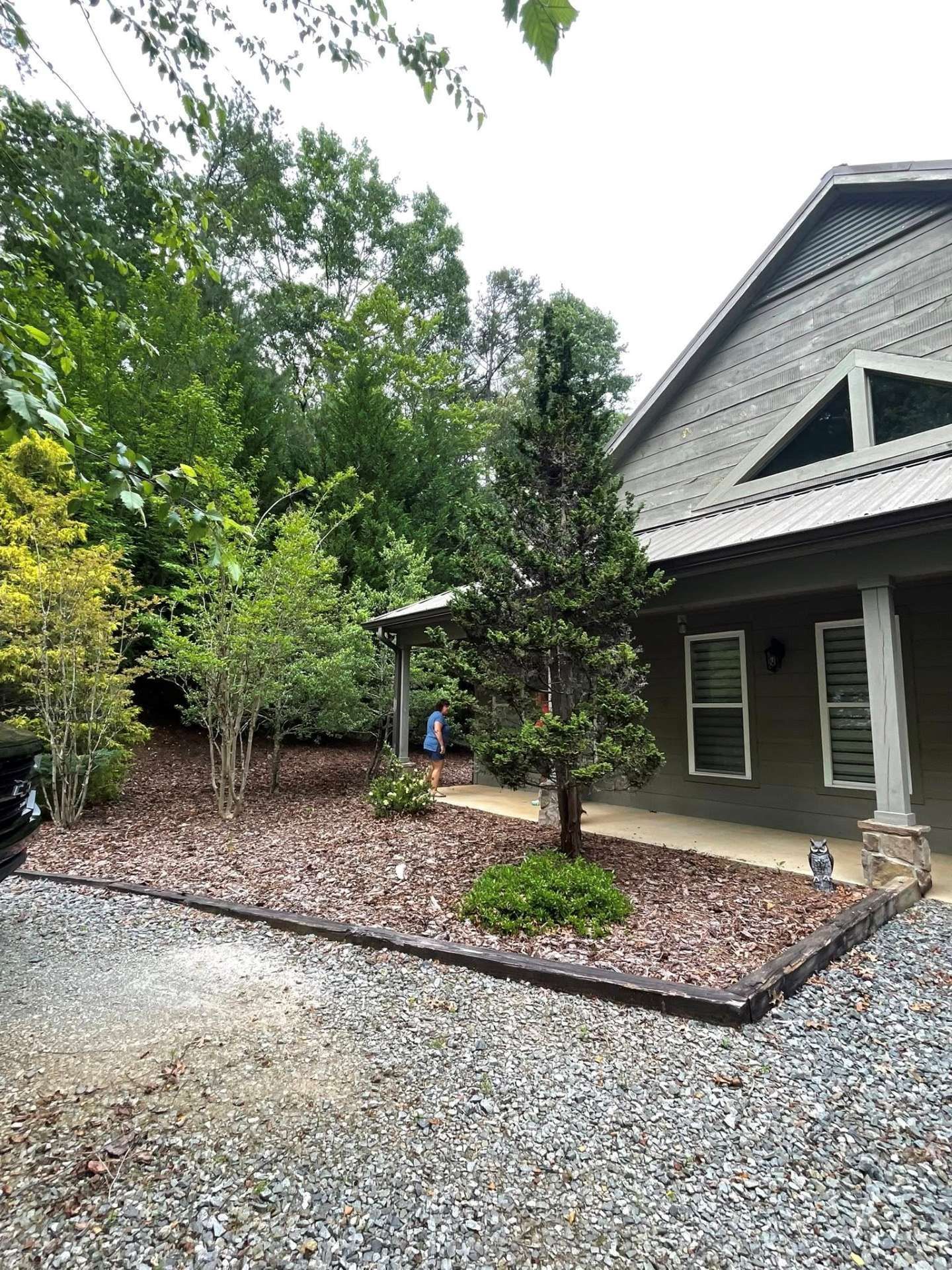 A gravel driveway leads to the covered porch of a gray house with dark wood trim, surrounded by trees and landscaping.