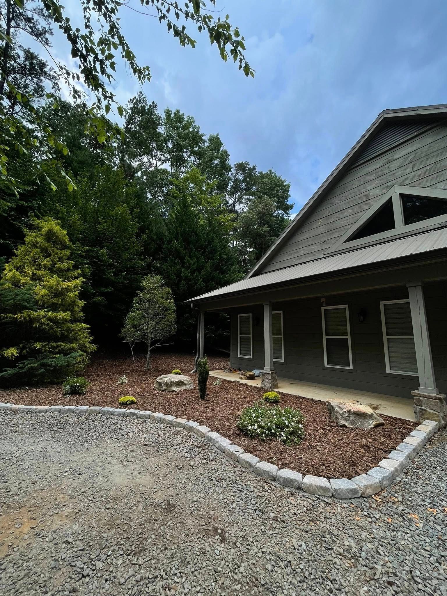 Gray house with a porch and stone-edged garden bed filled with mulch, shrubs, and trees under a cloudy sky.
