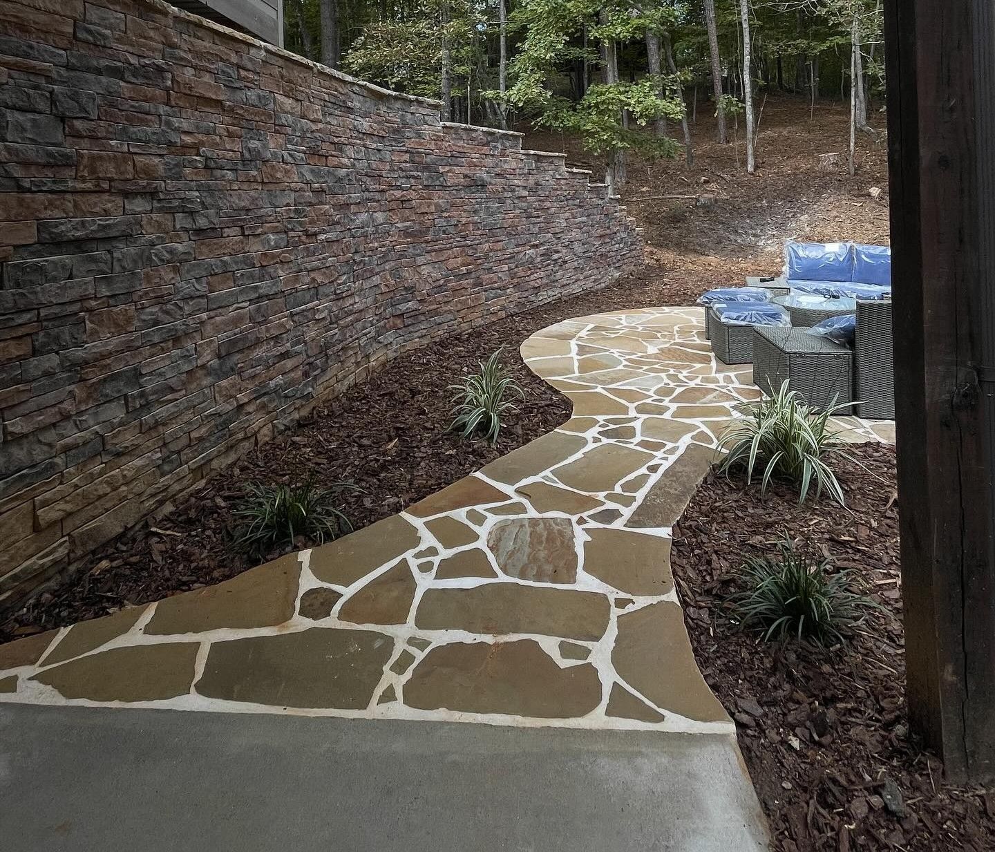 Flagstone pathway leading toward an outdoor seating area next to a stacked stone retaining wall.