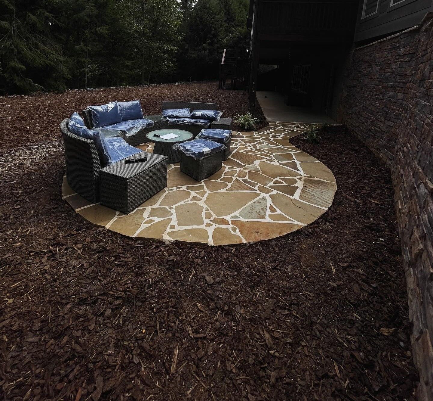 A curved stone patio with dark gray outdoor furniture sits in a mulched backyard next to a brick house.