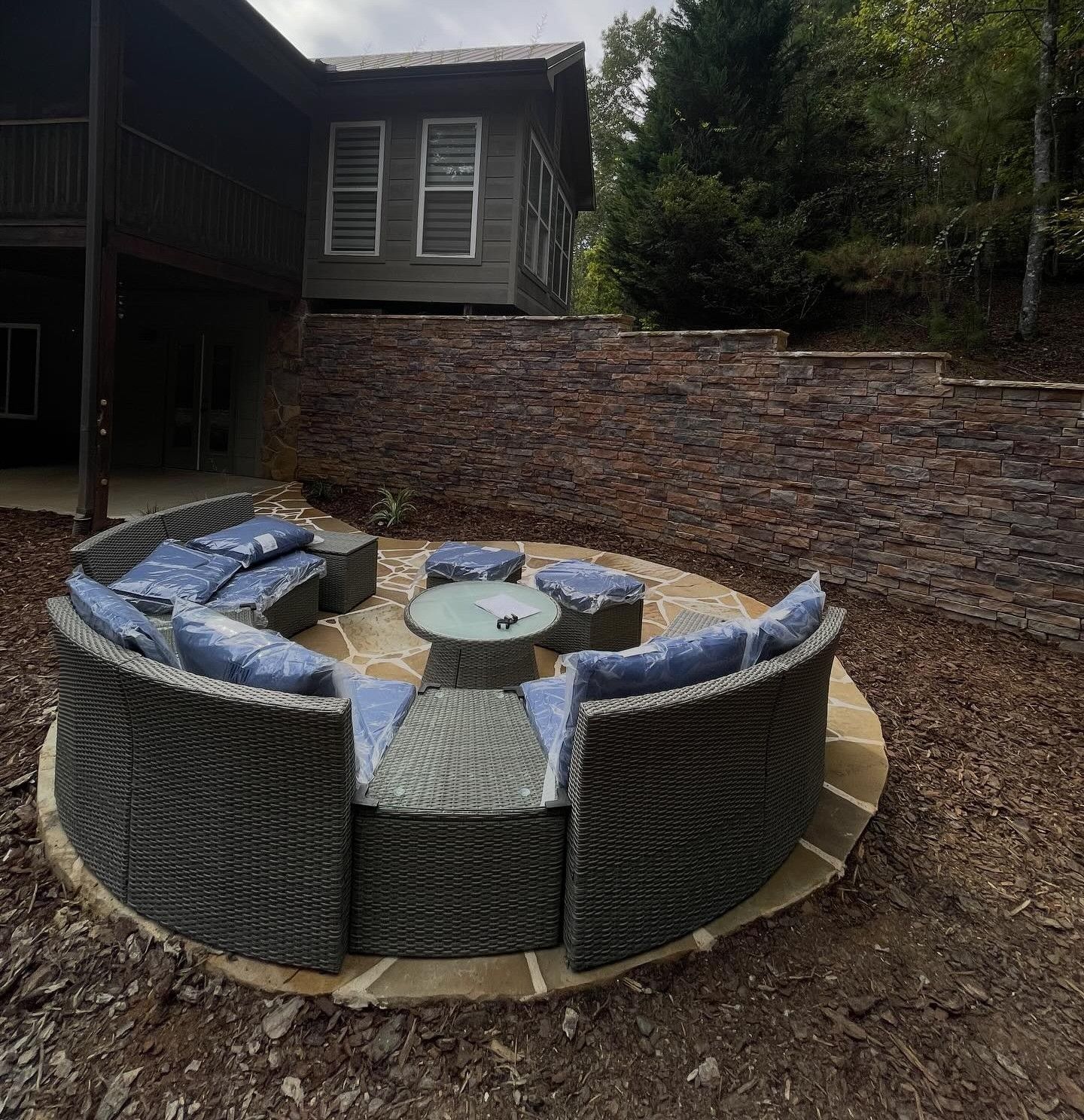 Curved wicker outdoor sofa and fire pit arranged on a stone patio in front of a house and stone retaining wall.