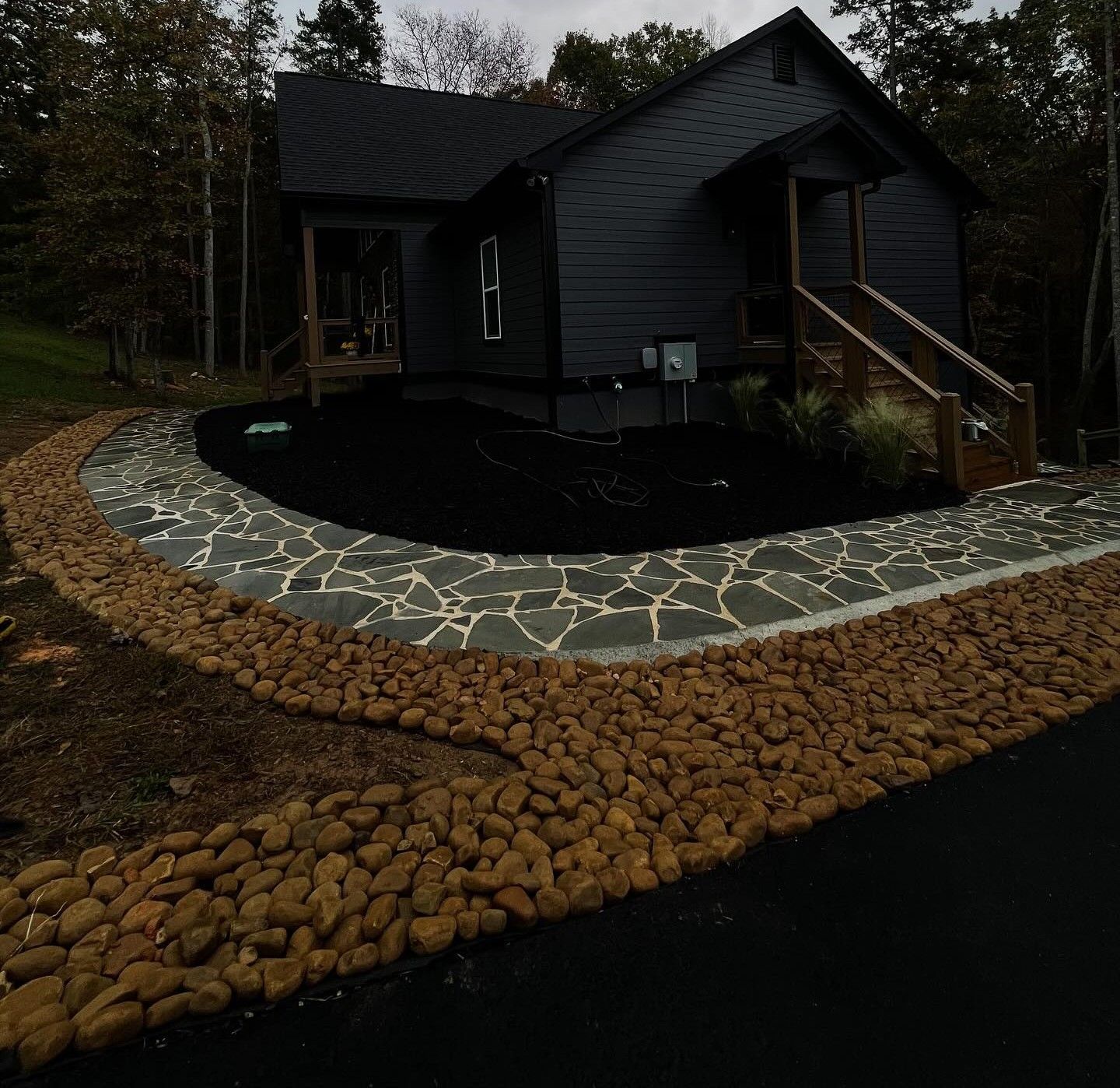 A dark house with a stone walkway bordered by brown river rocks, set against a backdrop of trees at dusk.