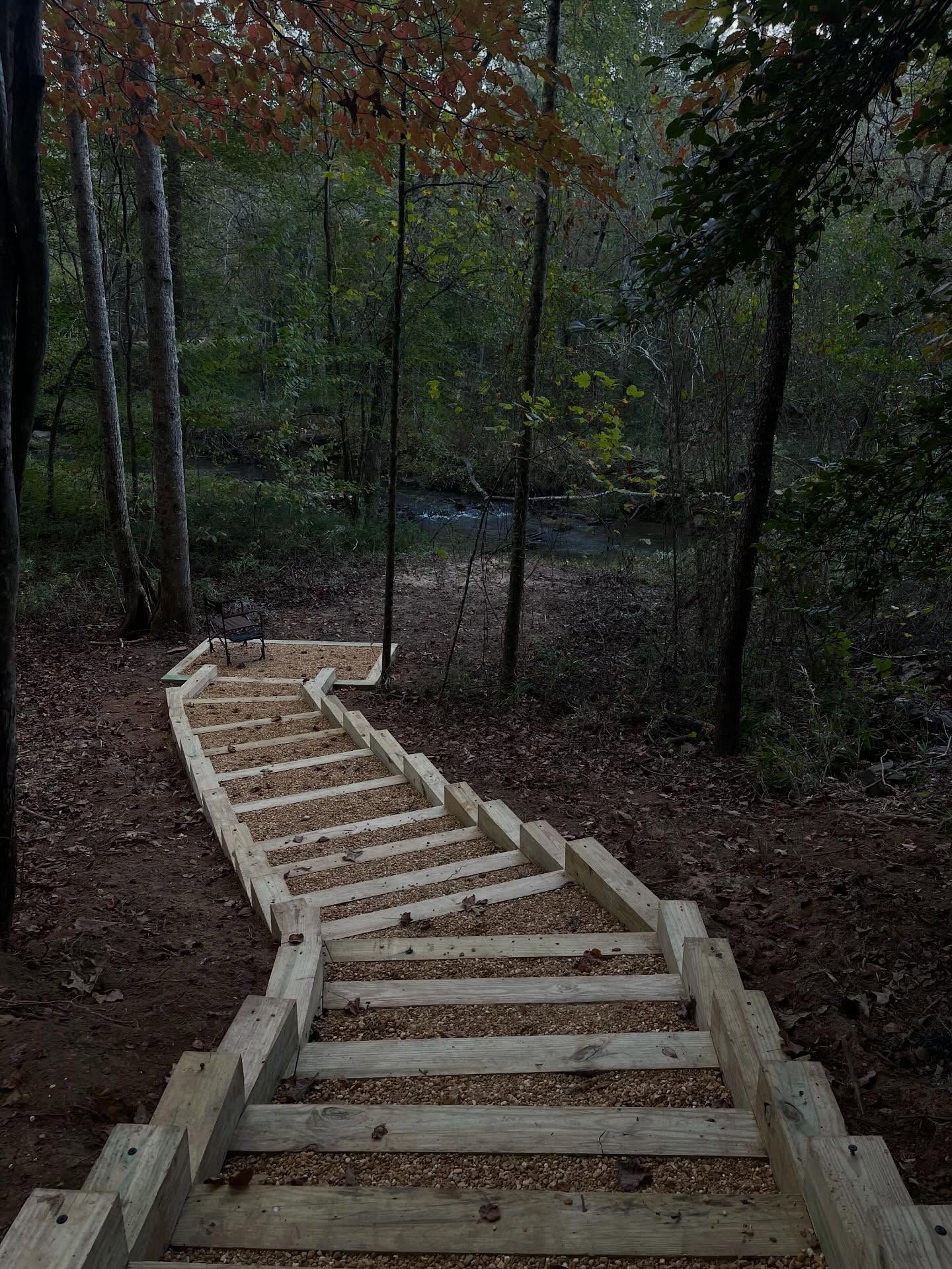 A set of wooden stairs filled with gravel winding down a wooded trail toward a stream in a forest setting.
