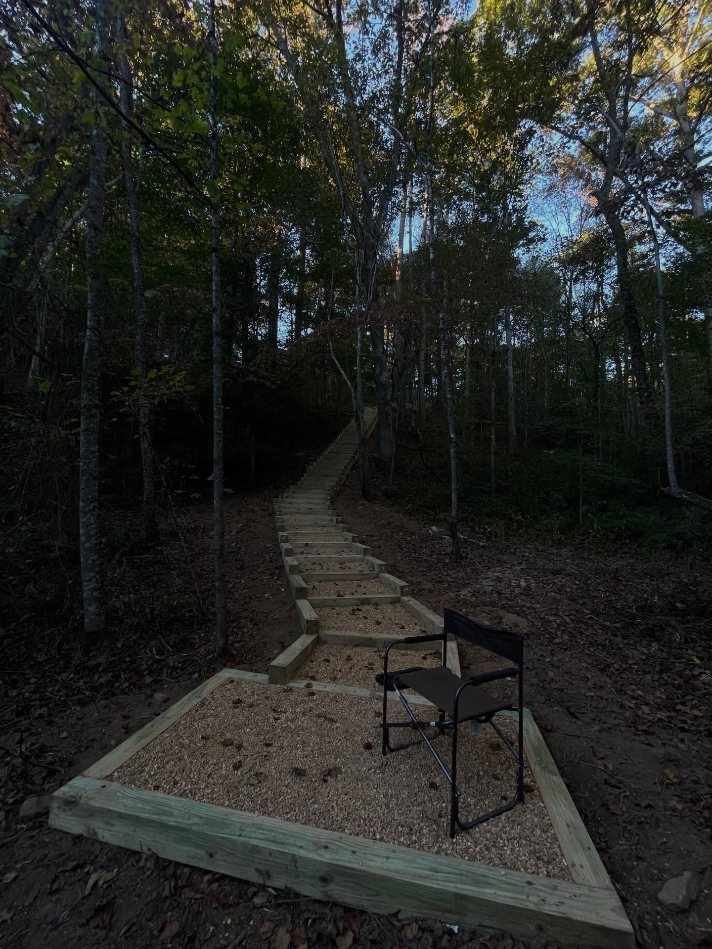A portable chair sits on a gravel landing at the base of a wooden-stepped trail winding through a forest.