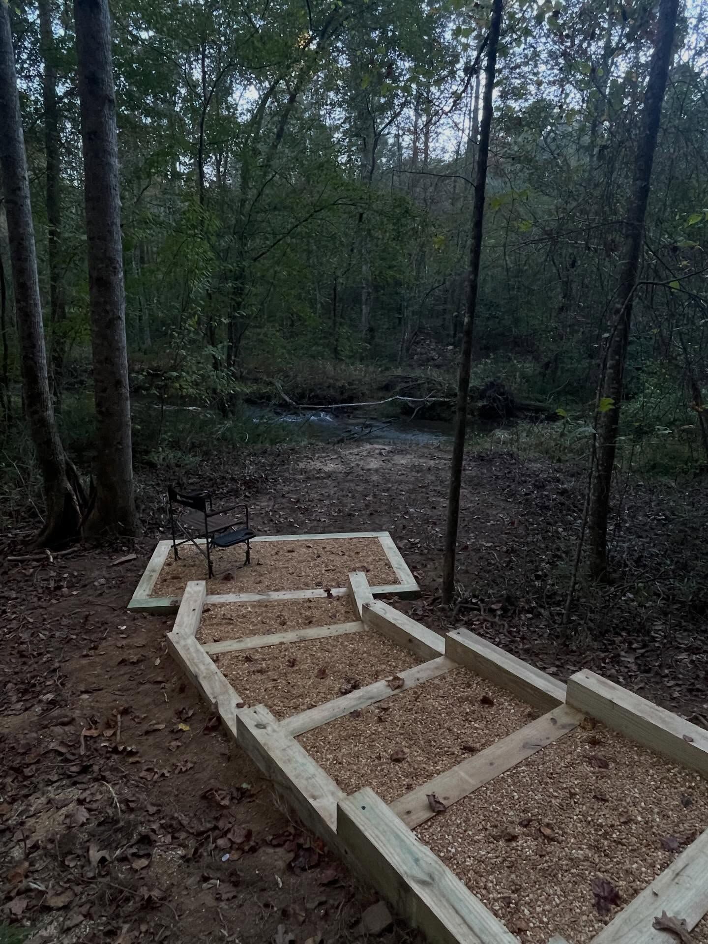 Wooden stairs filled with wood chips lead down a wooded trail toward a small bench overlooking a forest stream.