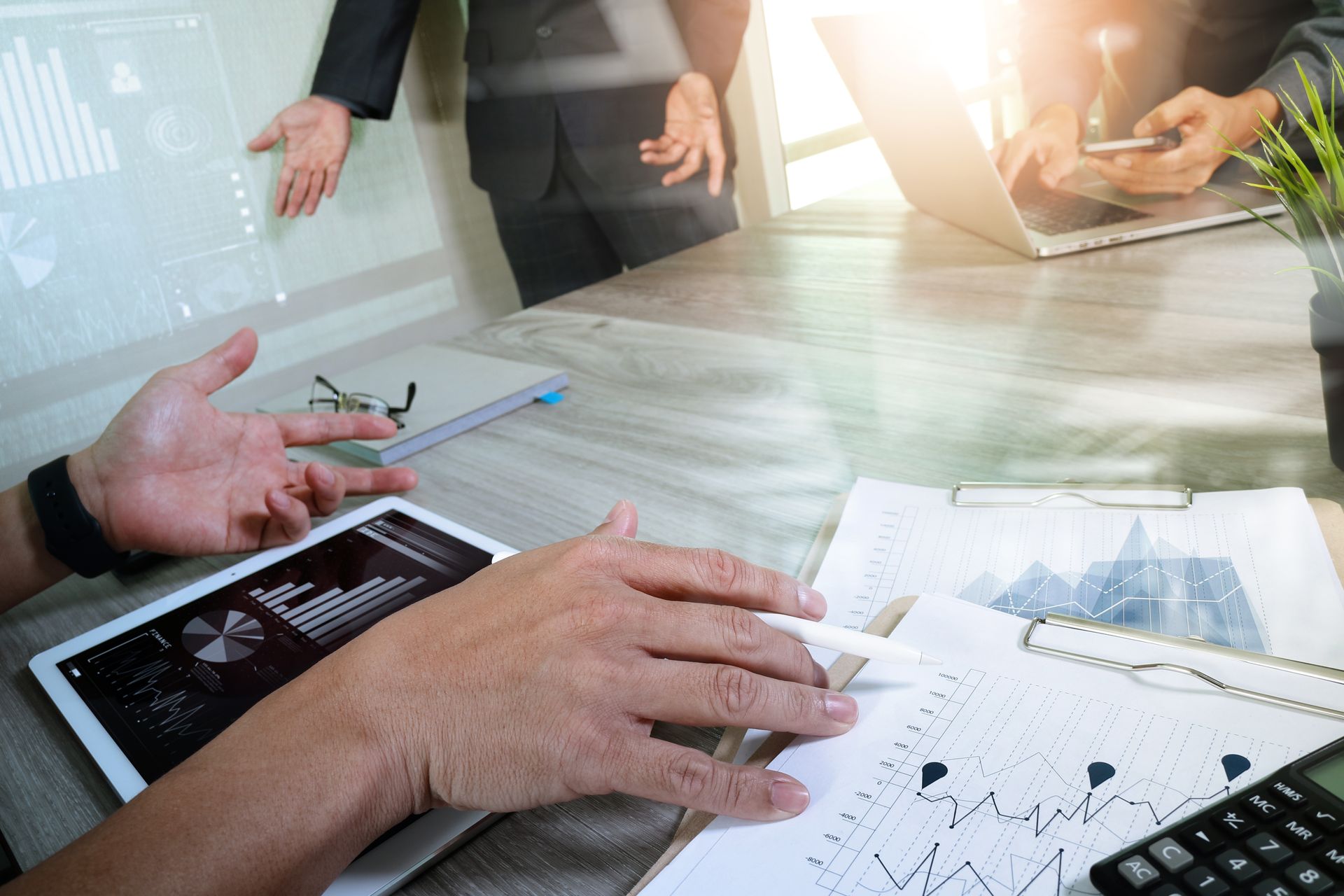 Business meeting at a table with charts, laptop, and hands gesturing in a bright office conference room