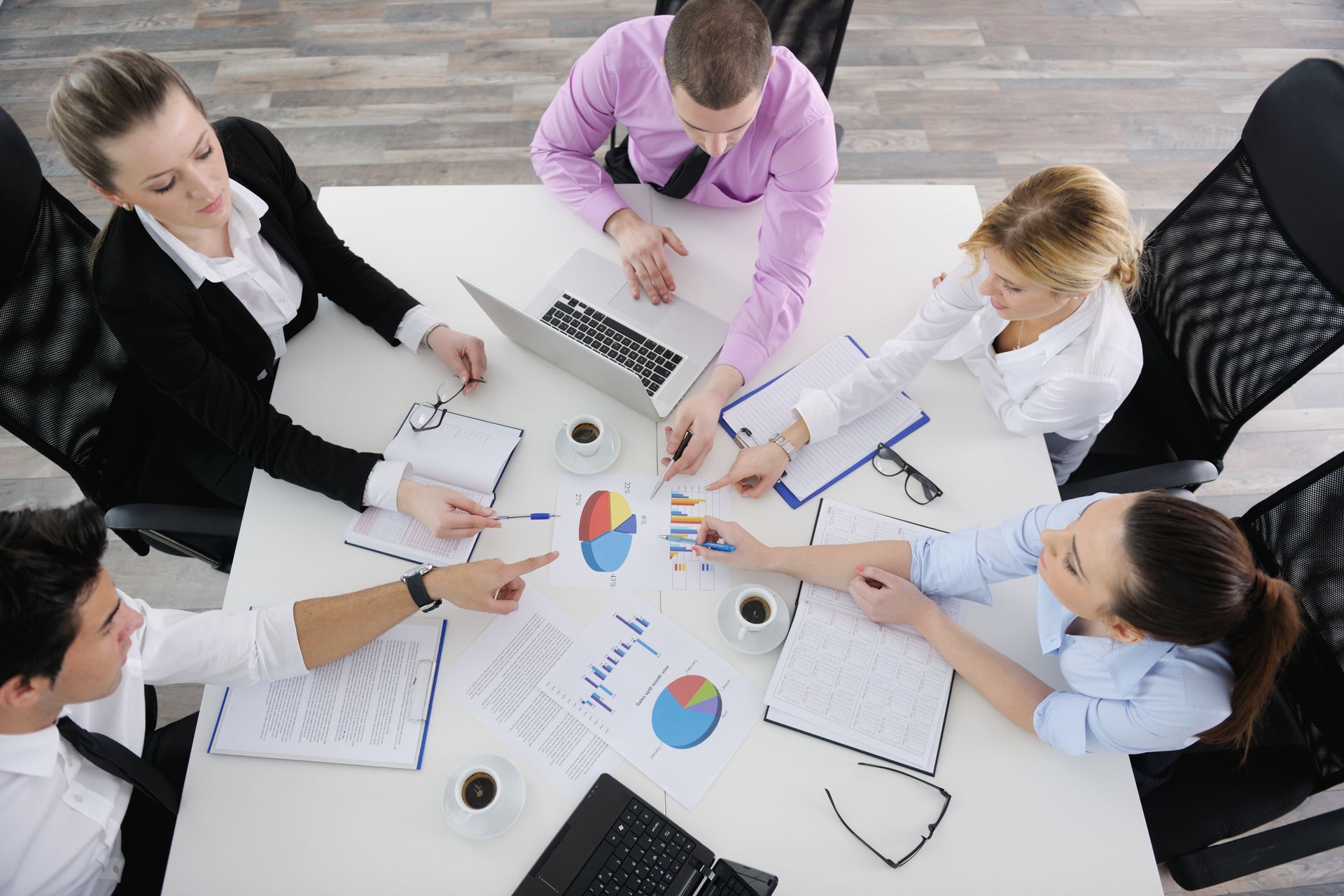 Team meeting around a white table with charts, laptops, and papers in a bright office