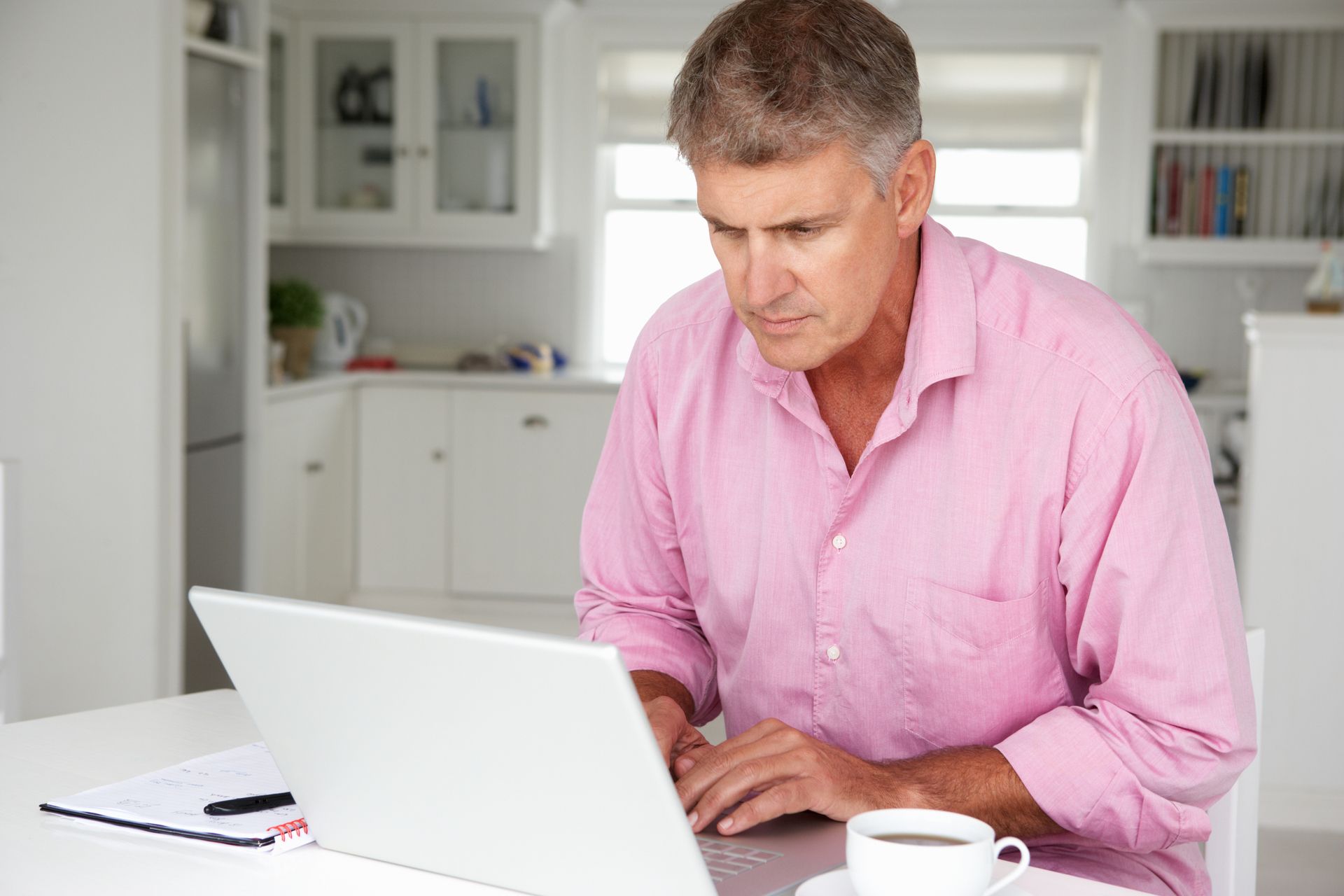 Man in pink shirt using a laptop at a white desk in a bright home office