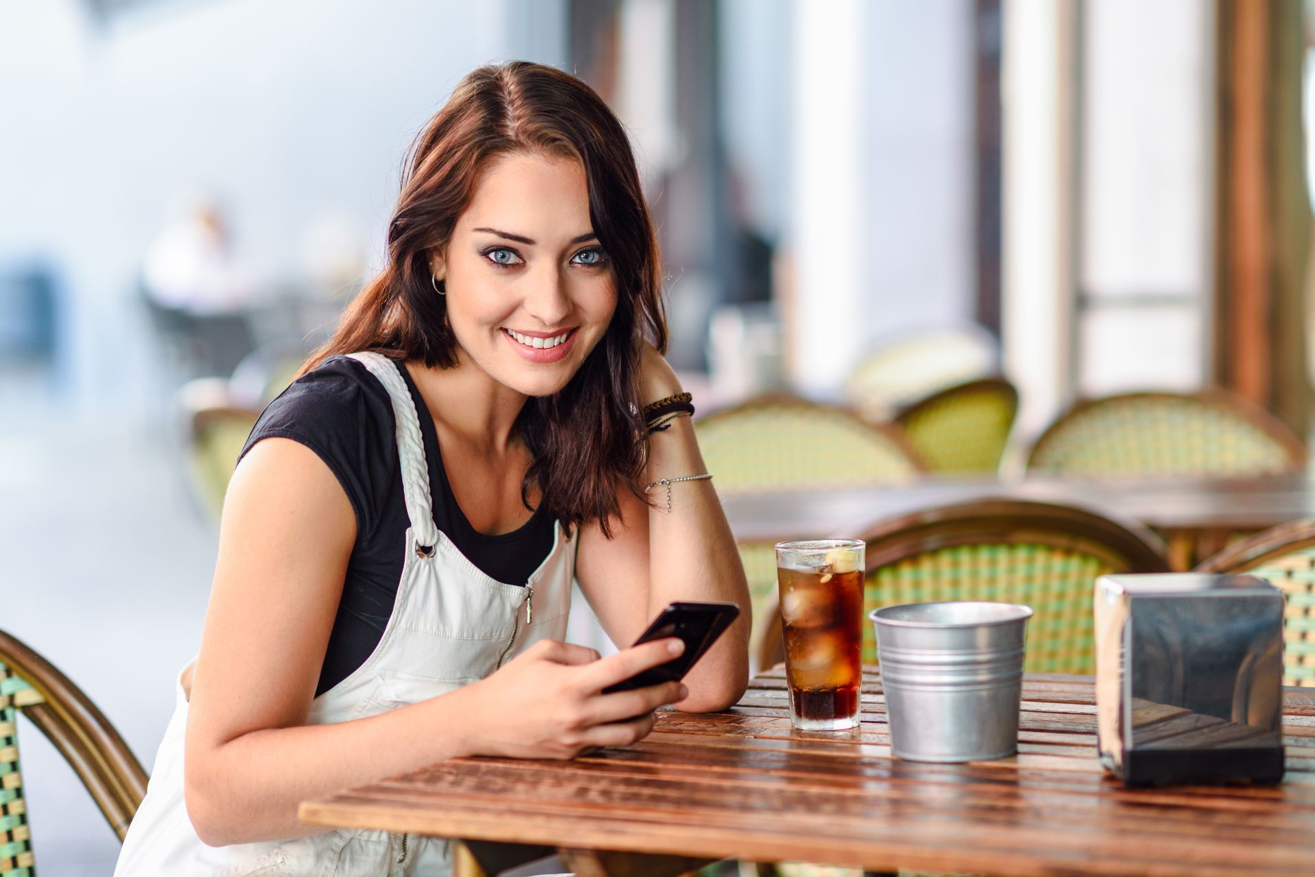 Woman smiling at a café table, holding a phone beside a drink and sugar container.