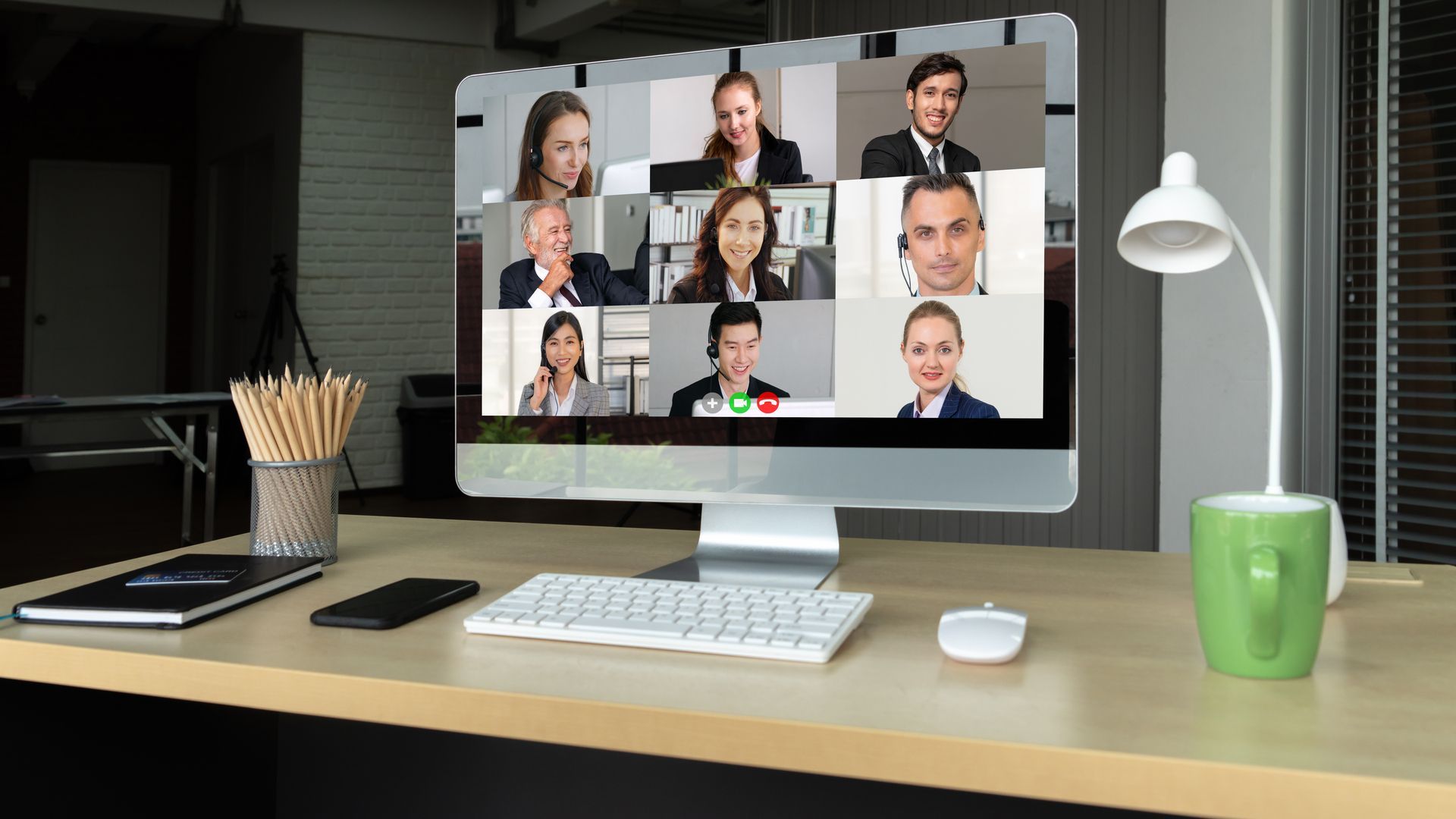 Desk with desktop showing a video conference grid, keyboard, lamp, and office supplies.