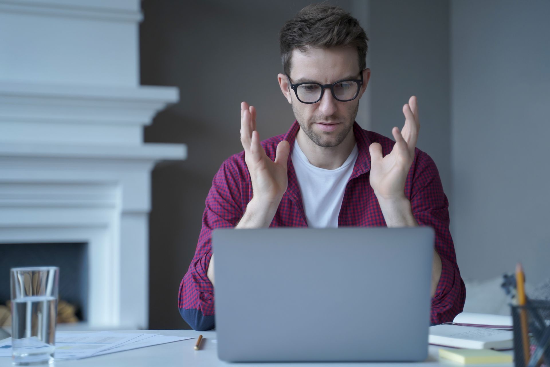 Man in glasses gesturing at laptop in a bright office, with papers, books, and a glass of water nearby