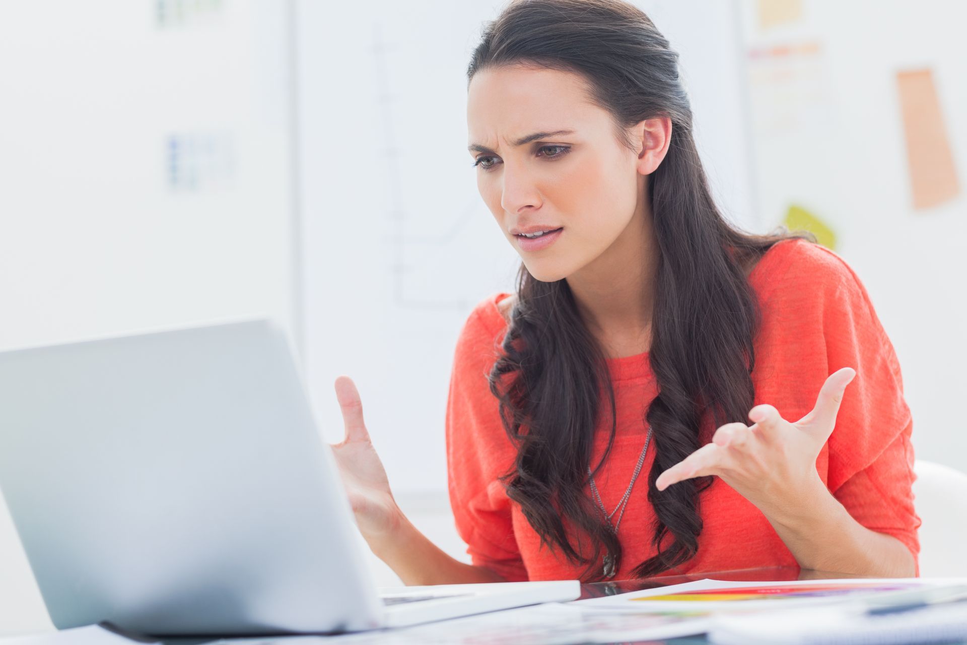 Woman in a coral top gesturing frustrated at a laptop in a bright office