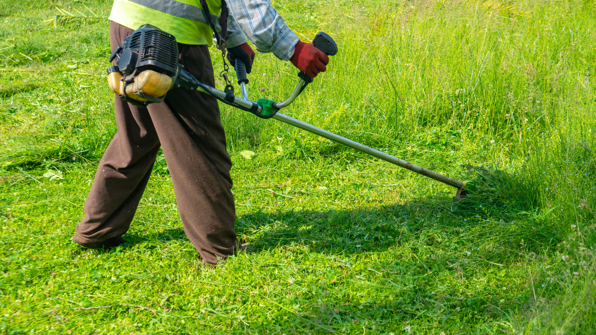Person using a string trimmer to cut tall grass in a green field, wearing a safety vest and gloves.