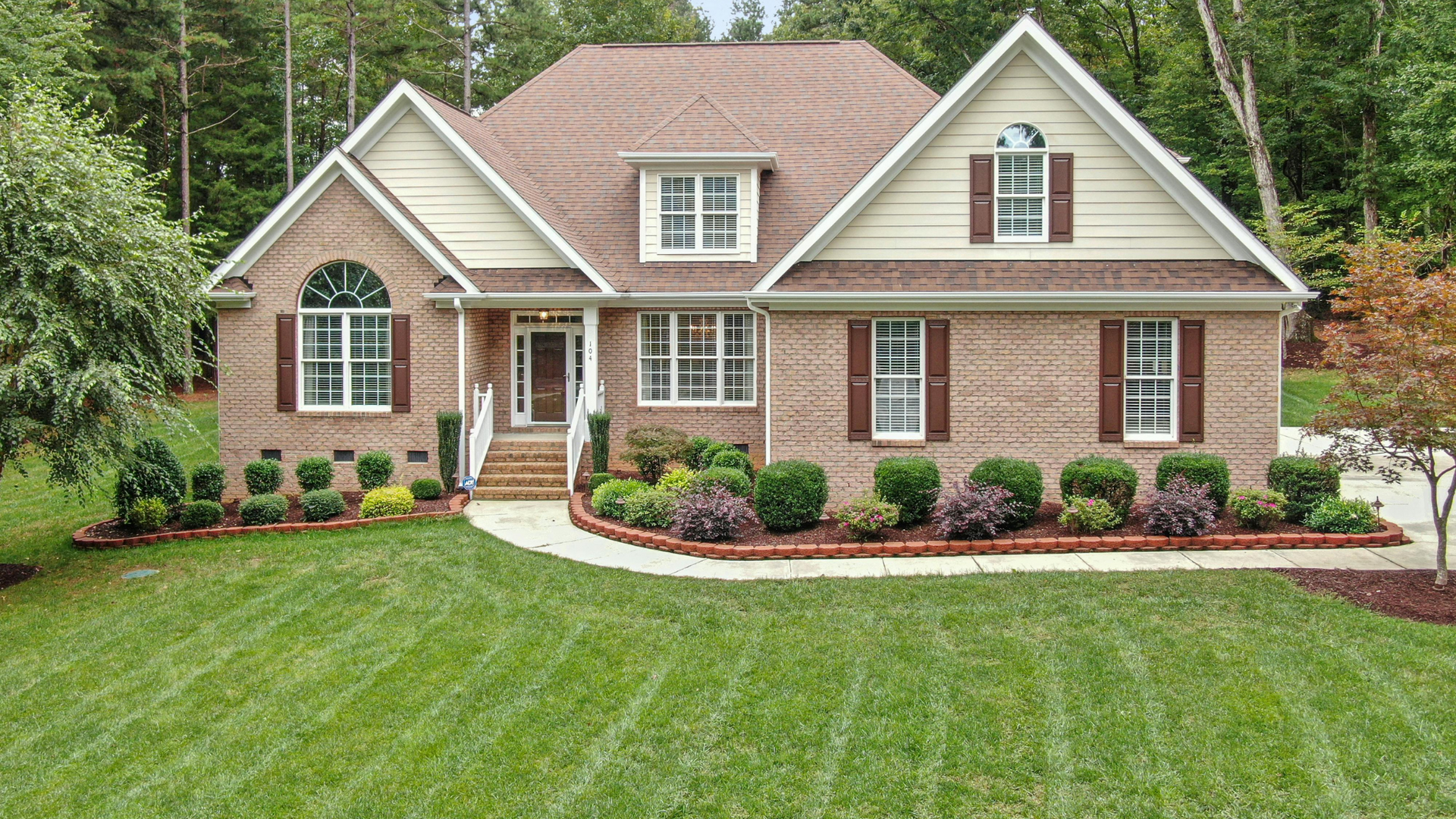 Brick house with brown roof, shutters, and landscaping, green lawn.