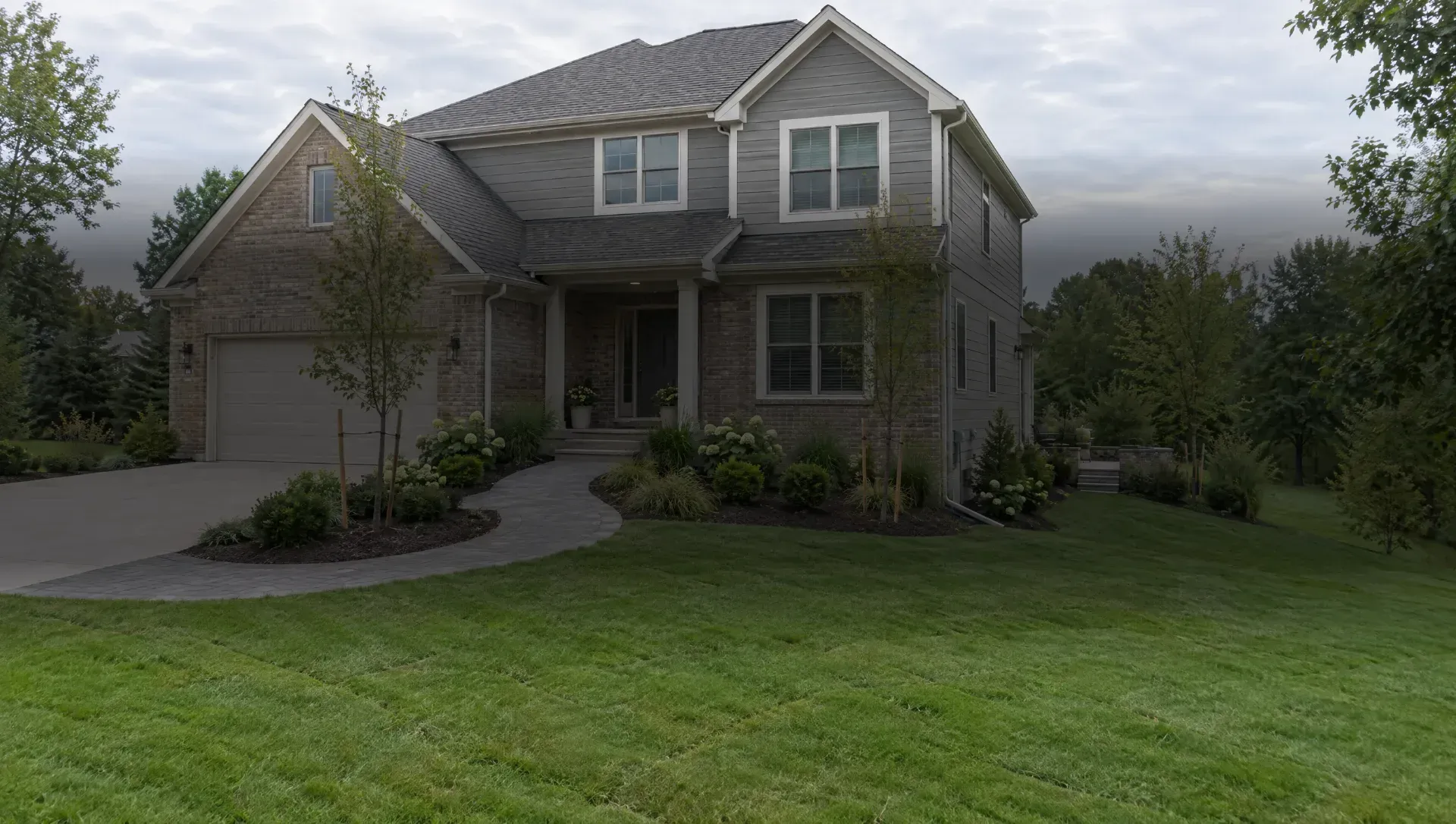 Illustration of a brick house with a curving paver walk, planting beds, two staked trees and a lawn.