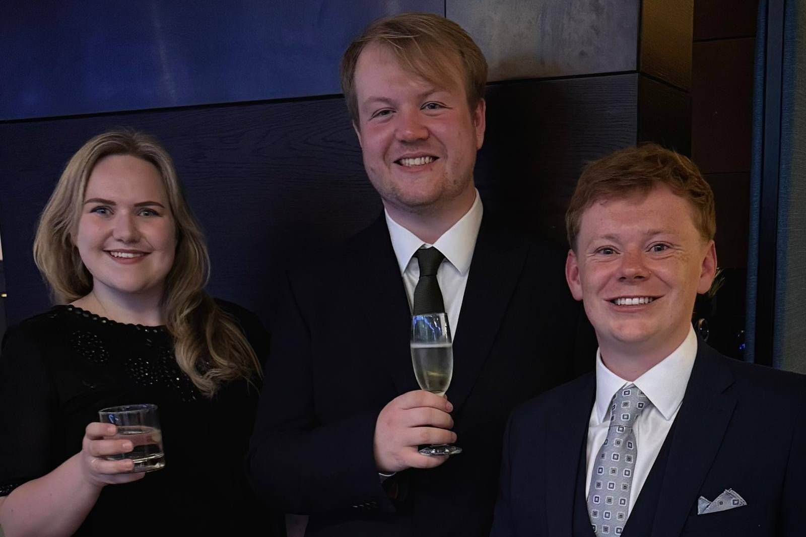 Three people in formalwear smiling and holding drinks at a dimly lit event.