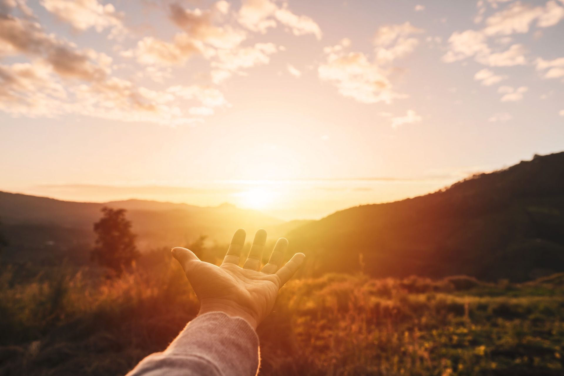 Hand reaching toward a golden sunset over a grassy valley and hills