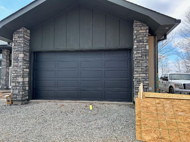 Dark gray garage door with stone pillars. A white truck is parked nearby. Modern house exterior with a dark garage door, stone pillars, gravel driveway, and a white van on the right.
