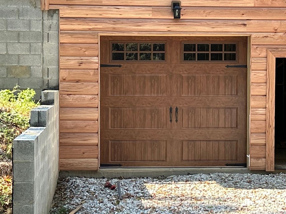 Wooden garage door with windowed top panels, set in wood siding. Wooden garage door with small windows and side entrance on a gravel driveway