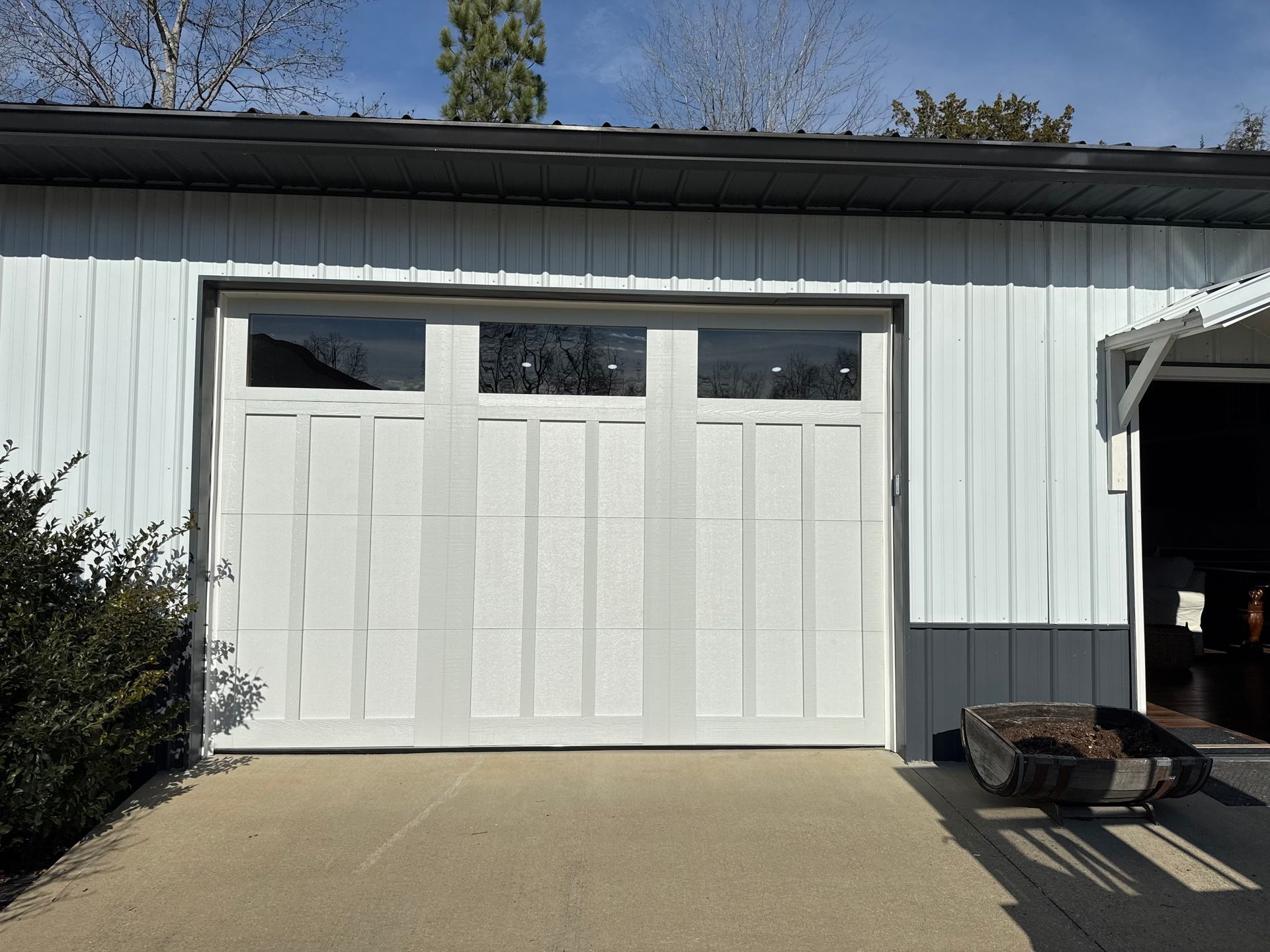 White garage door with glass panels on a building with white and gray siding. White garage door on a gray house with concrete driveway and a trash bin at right