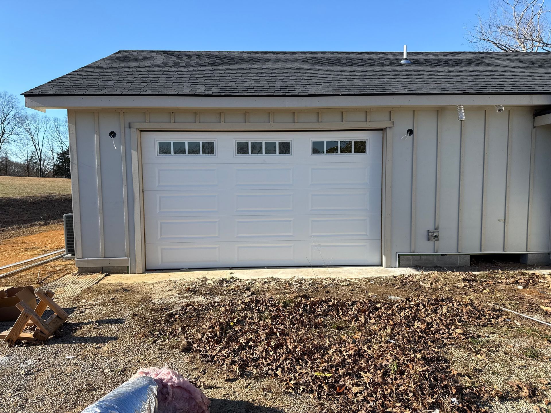 White garage door on a light-colored building under a dark roof, surrounded by brown leaves and dirt. White garage door on a beige house with dark shingle roof, seen from a yard in daylight