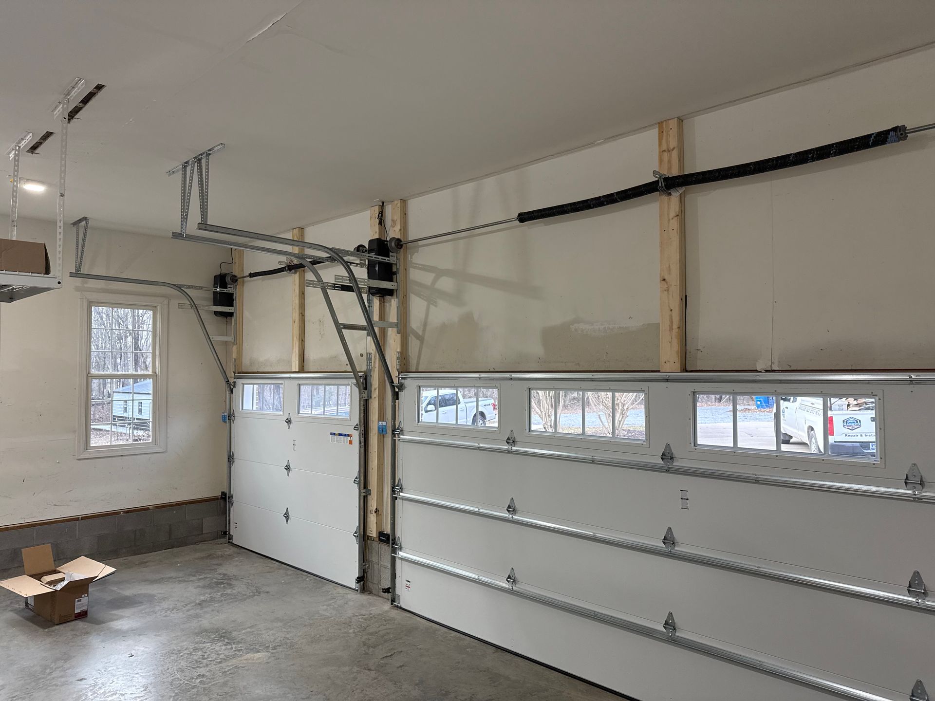 Garage interior with two white garage doors and an automated opening system. Empty garage interior with white walls, concrete floor, windows, and a large closed garage door.