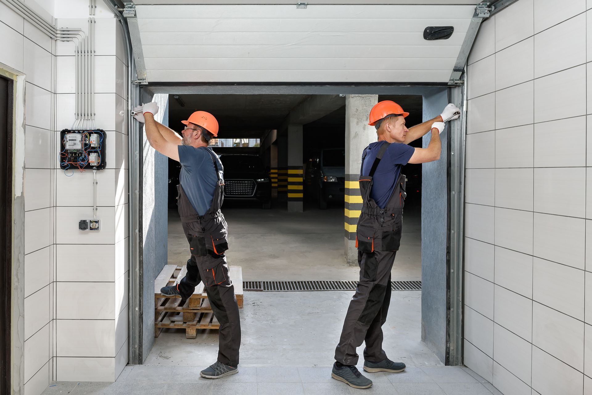 Two workers in safety gear installing a garage door. Two workers in orange hard hats operating a large roll-up door in a tiled industrial hallway