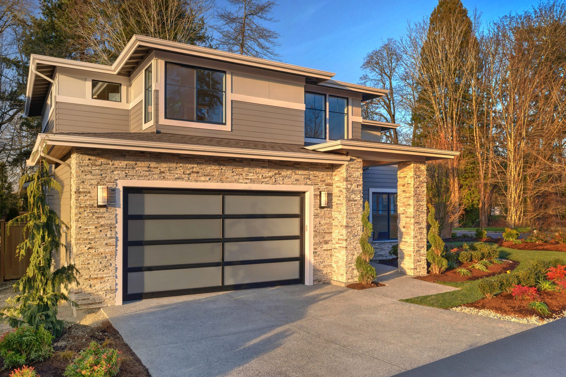 Modern two-story house with stone facade and gray garage door, set in a yard with landscaping.