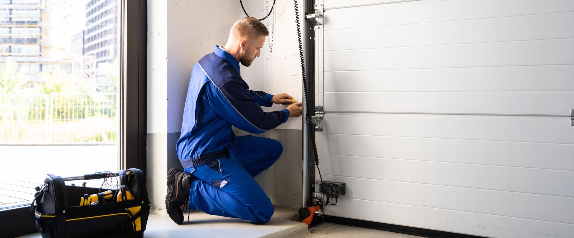 Landscaped flower bed with mixed colorful plants and green lawn edge. Technician in blue coveralls kneels beside a garage door track, adjusting hardware with a tool.