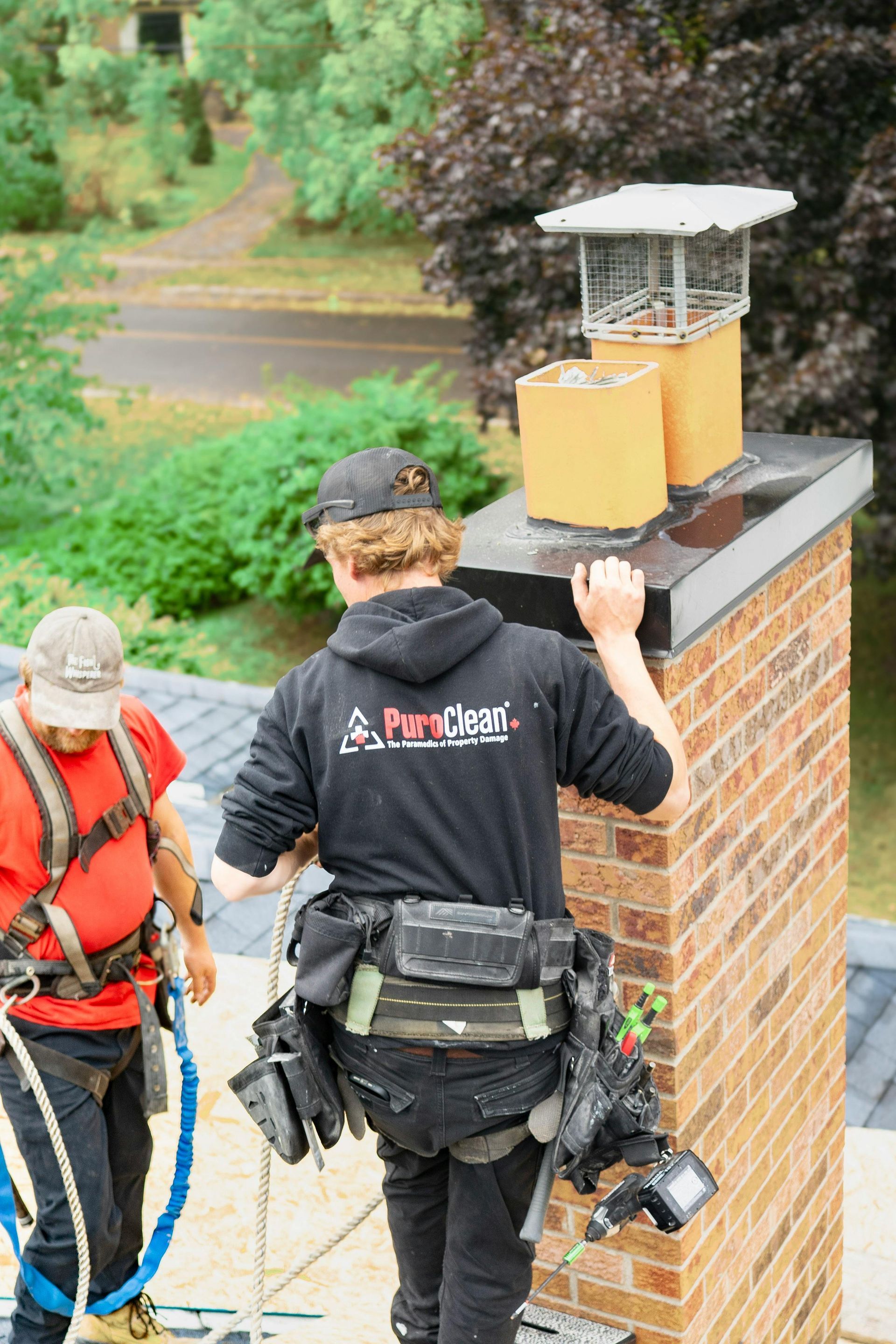 Two people on a roof working on a chimney. One in a safety harness and the other is holding onto the top of the chimney.
