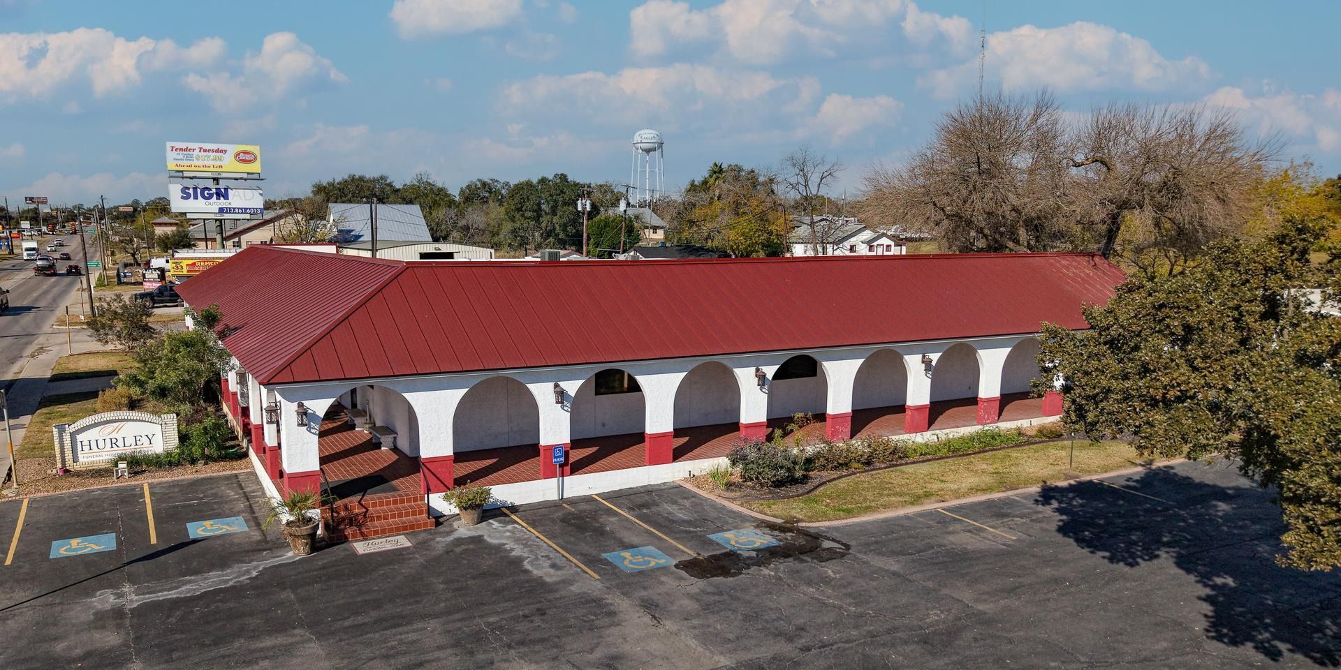Aerial view of a red-roofed building with white arches and parking lot, surrounded by trees under a blue sky.