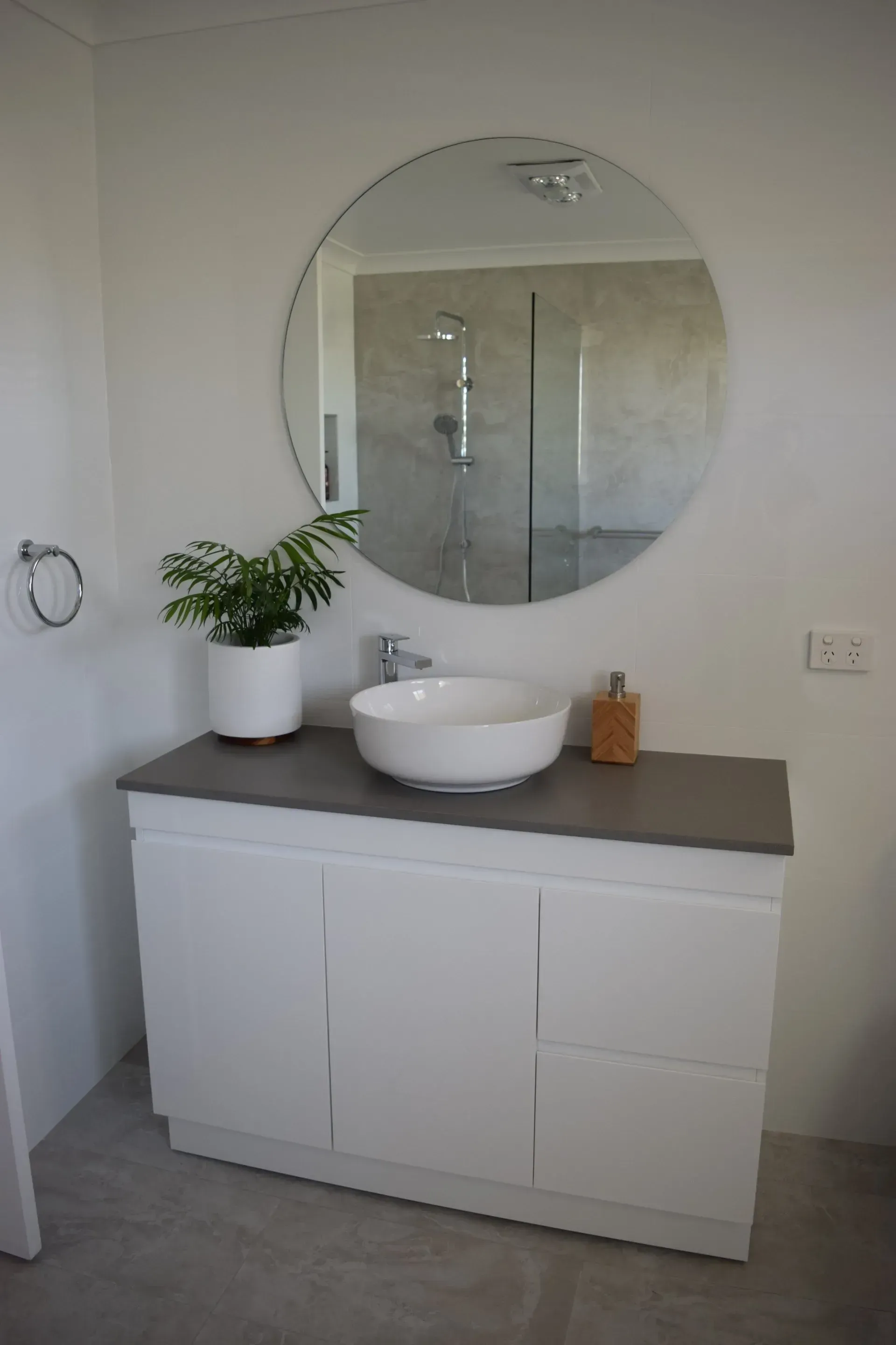 White bathroom vanity with round mirror, sink, and plant; gray countertop and wall.