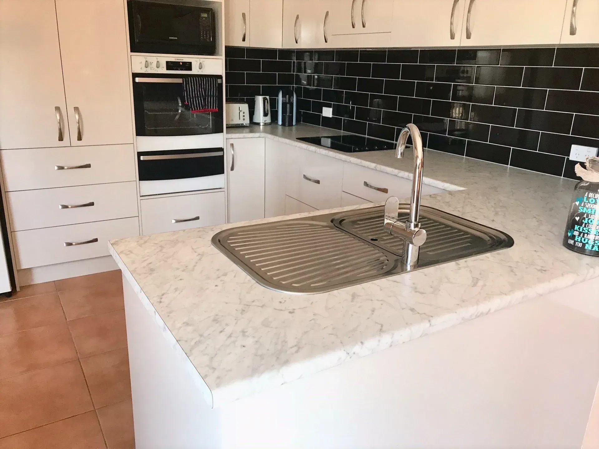 White kitchen with black tiled backsplash, stainless steel sink, and built-in appliances.