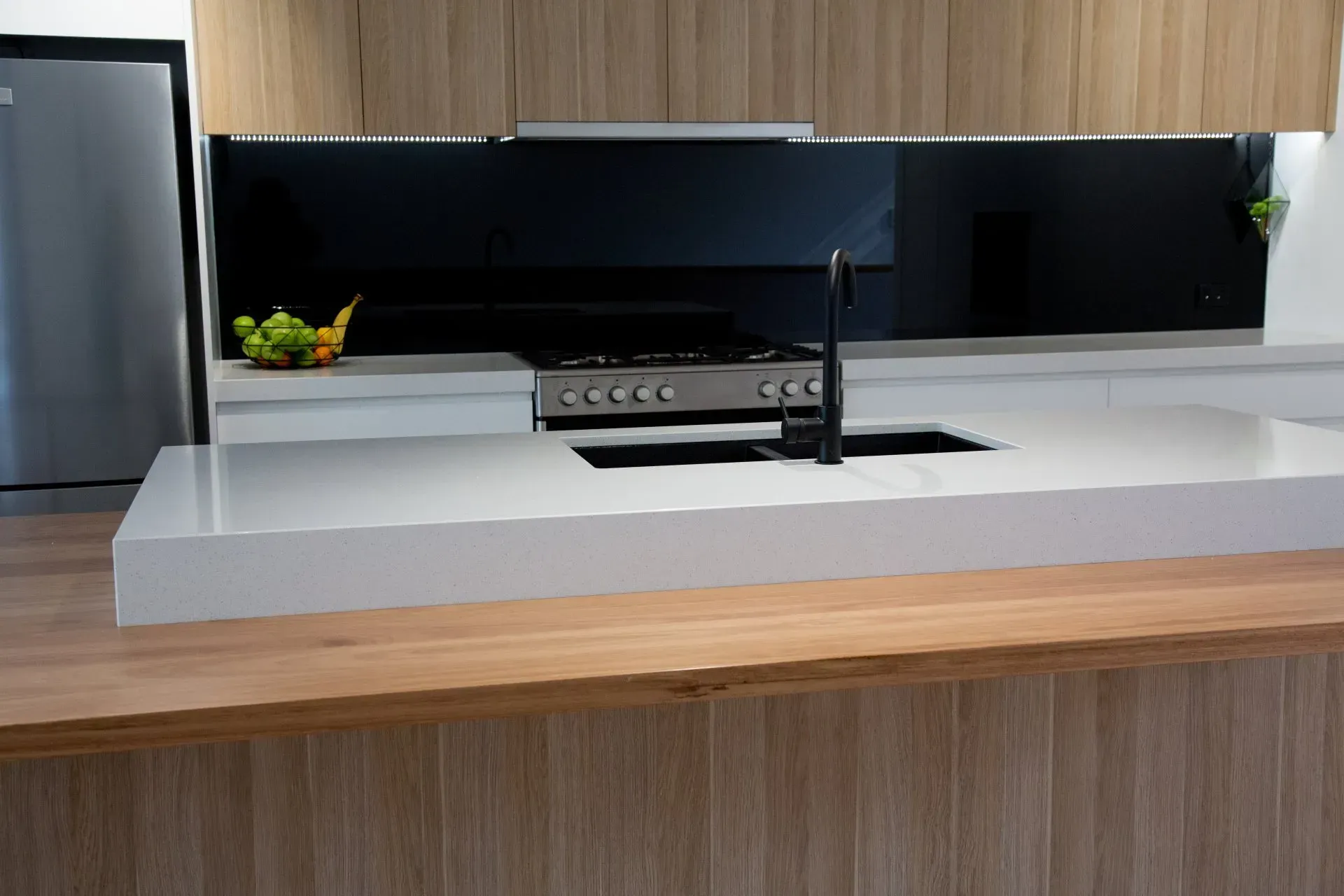 Kitchen with white countertop, black backsplash, stainless steel stove and sink. Wooden cabinets.