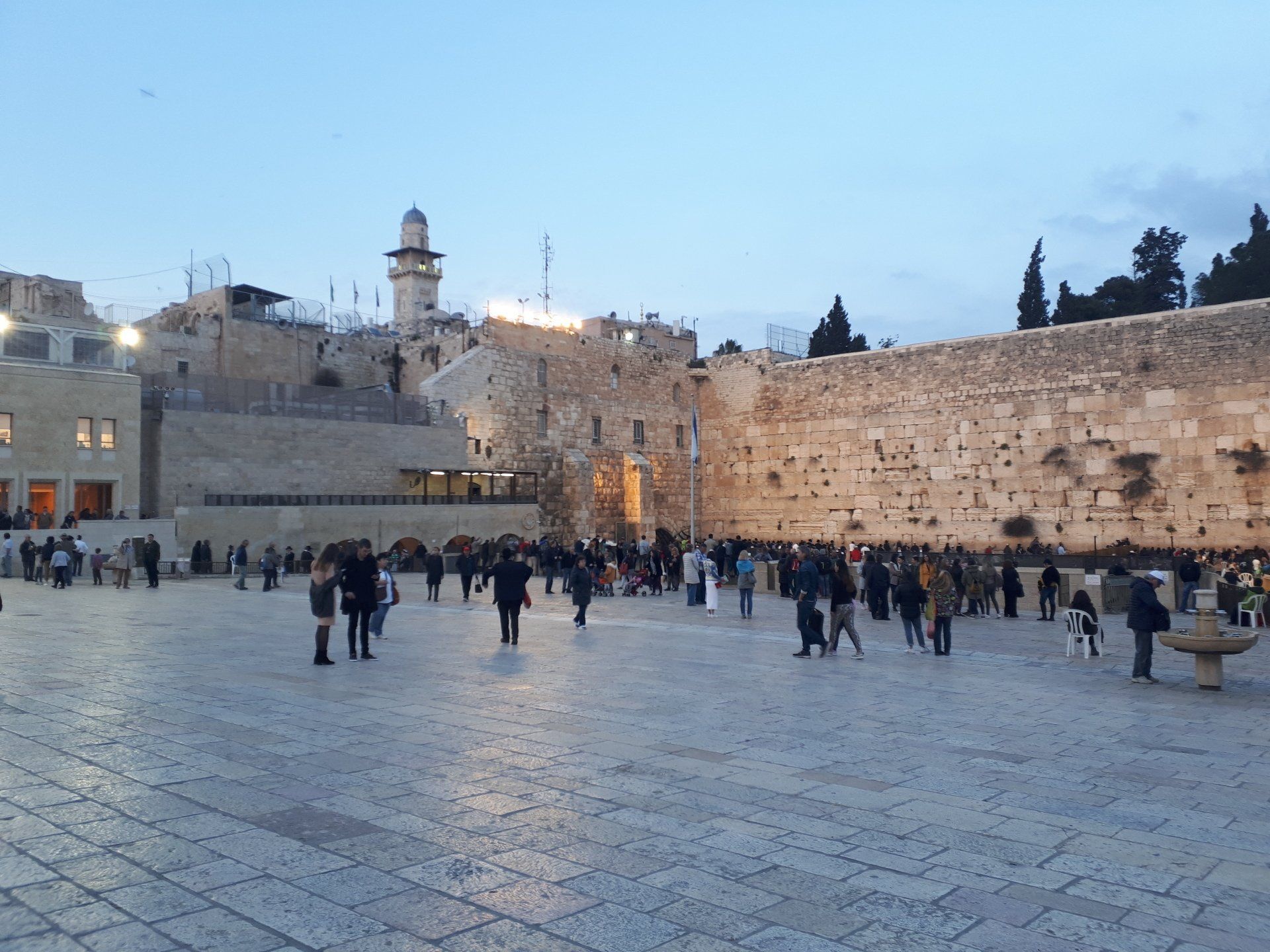 Western Wall plaza with people, building in background, minaret on the left.