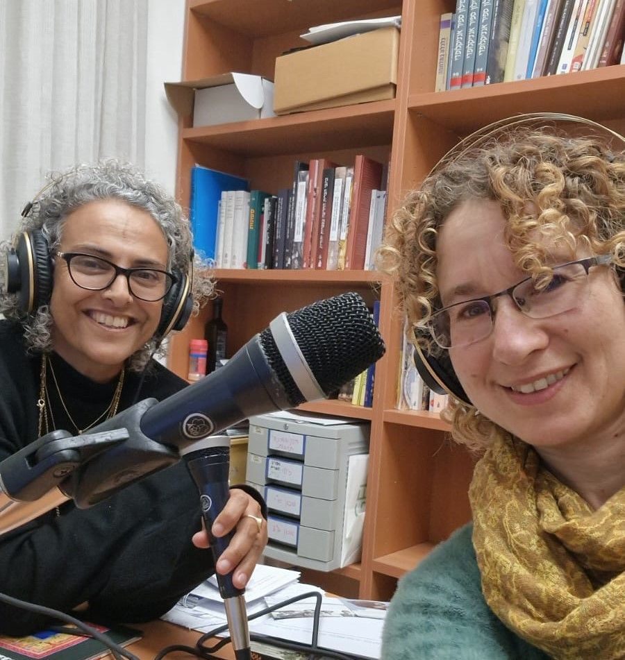 Two women in headphones at a microphone, smiling in a room with bookshelves.