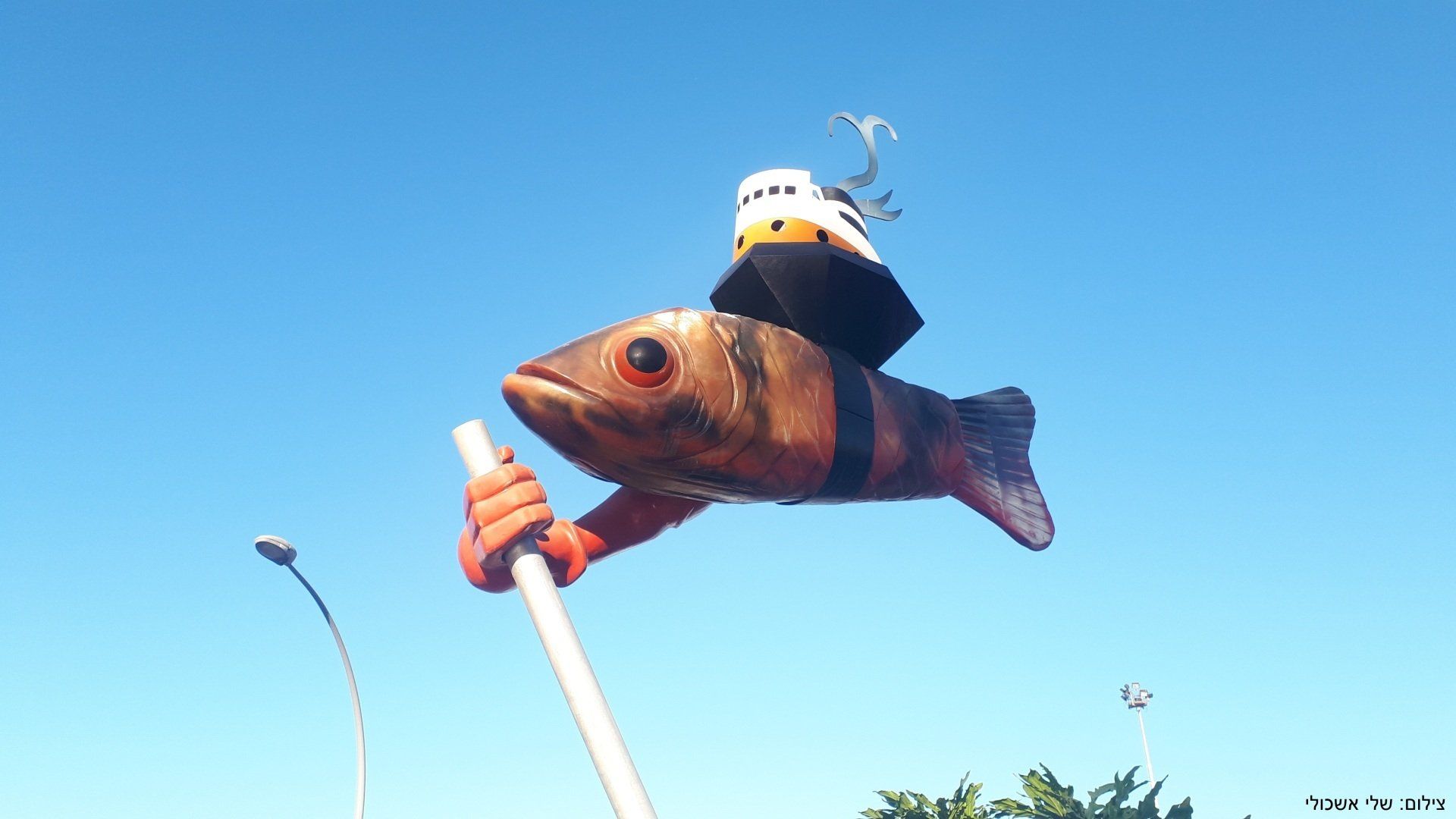 A large fish with an orange and black striped body holds a white pole, topped by a miniature ship under a blue sky.