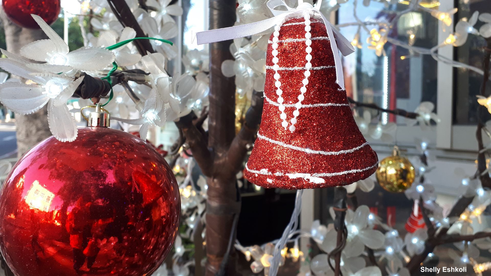 Red glitter Christmas ornaments, a bell, and a bauble hanging on a white-lit tree.