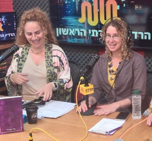 Two women smiling, seated at a radio studio table. One wears patterned jacket, other, a brown coat. Microphones, notes, and a coffee cup visible.
