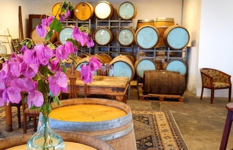 Purple flowers in a glass vase in front of a wine barrel; other barrels on a shelf in a rustic room.
